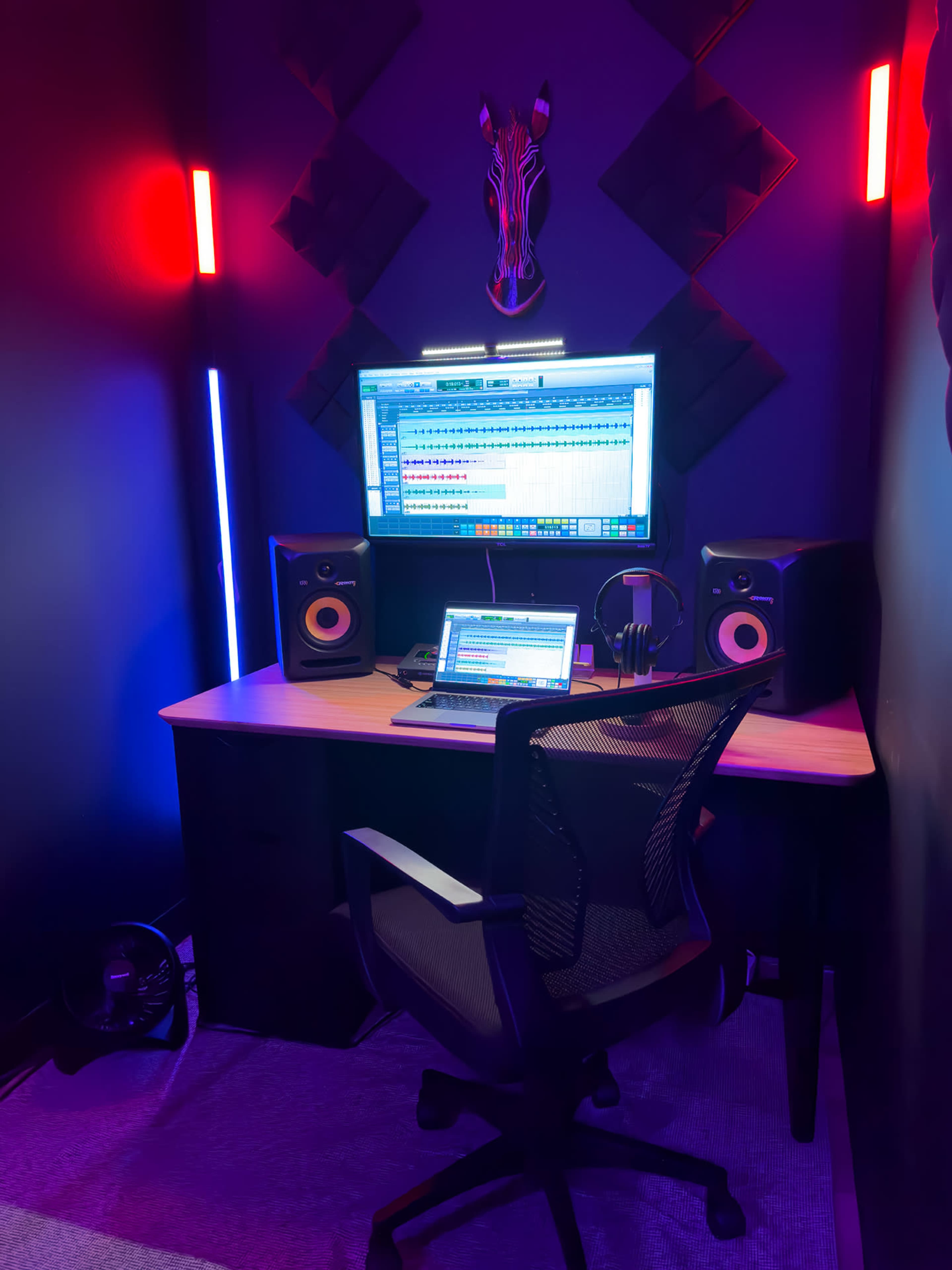The image shows a home music production studio featuring a desk with a computer monitor, speakers, a laptop, headphones, and colorful LED strip lighting on the walls.