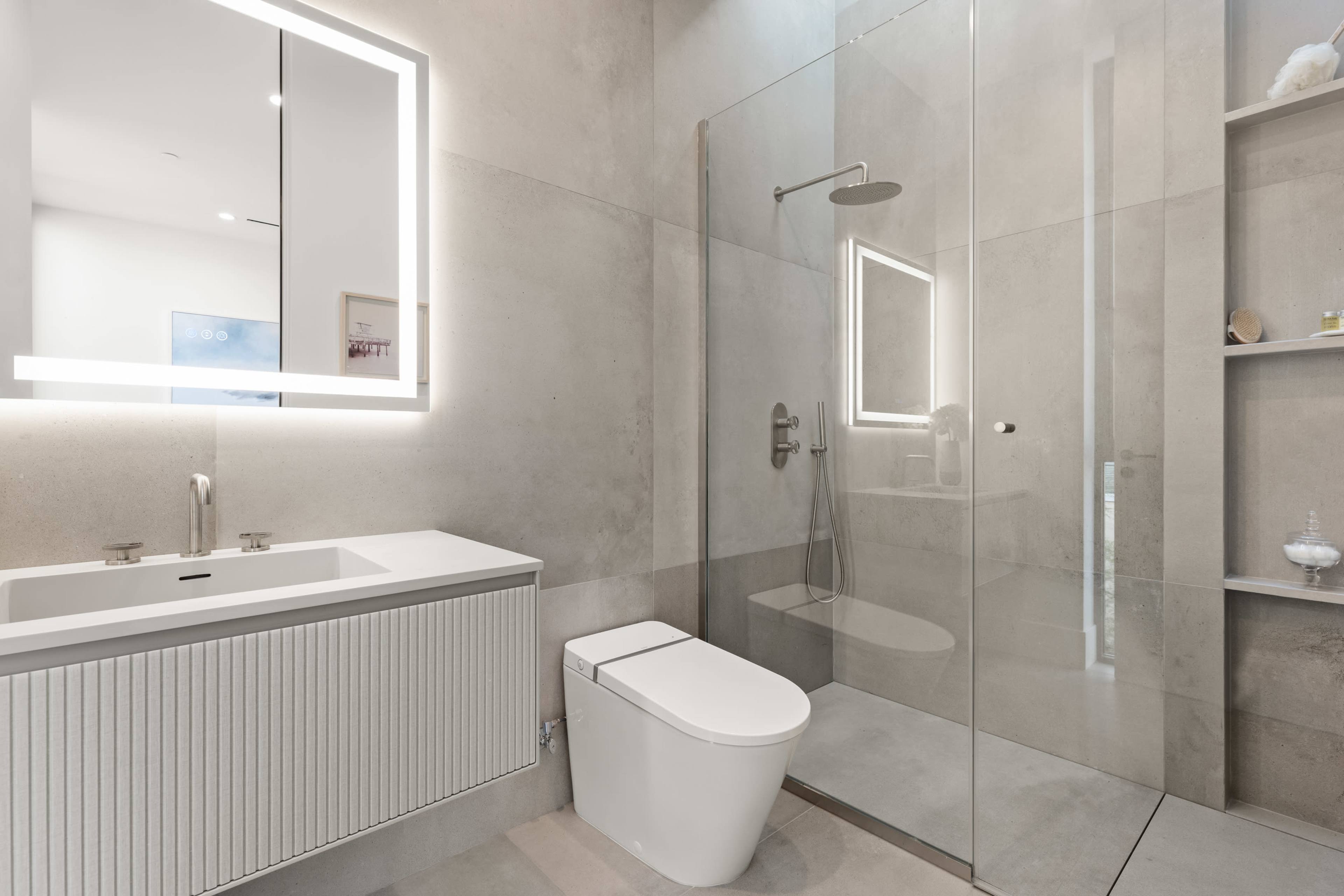 A modern bathroom with a glass shower, a wall-mounted sink, a sleek toilet, and a backlit mirror.