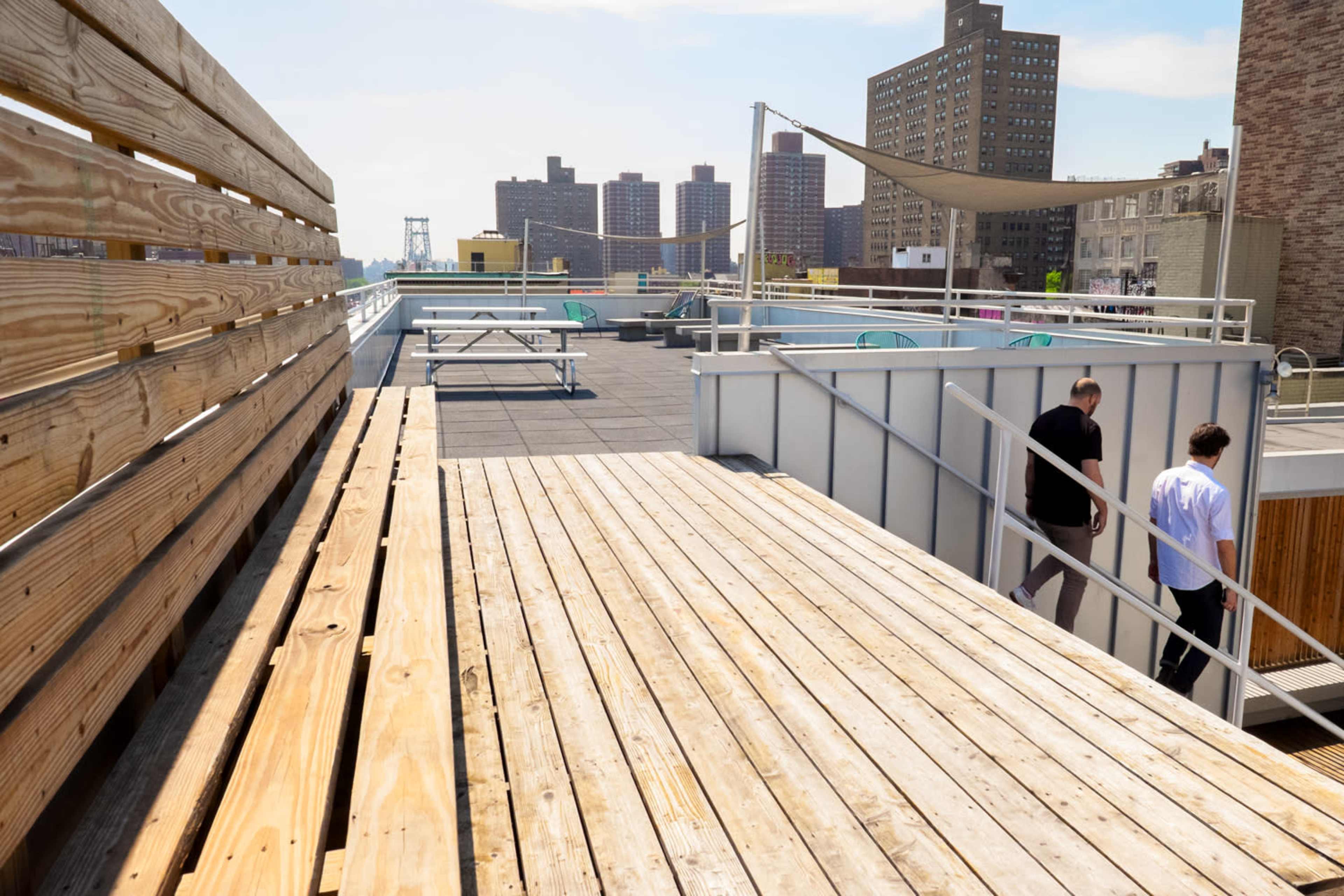 Two people walk along a wooden deck on a rooftop surrounded by buildings.