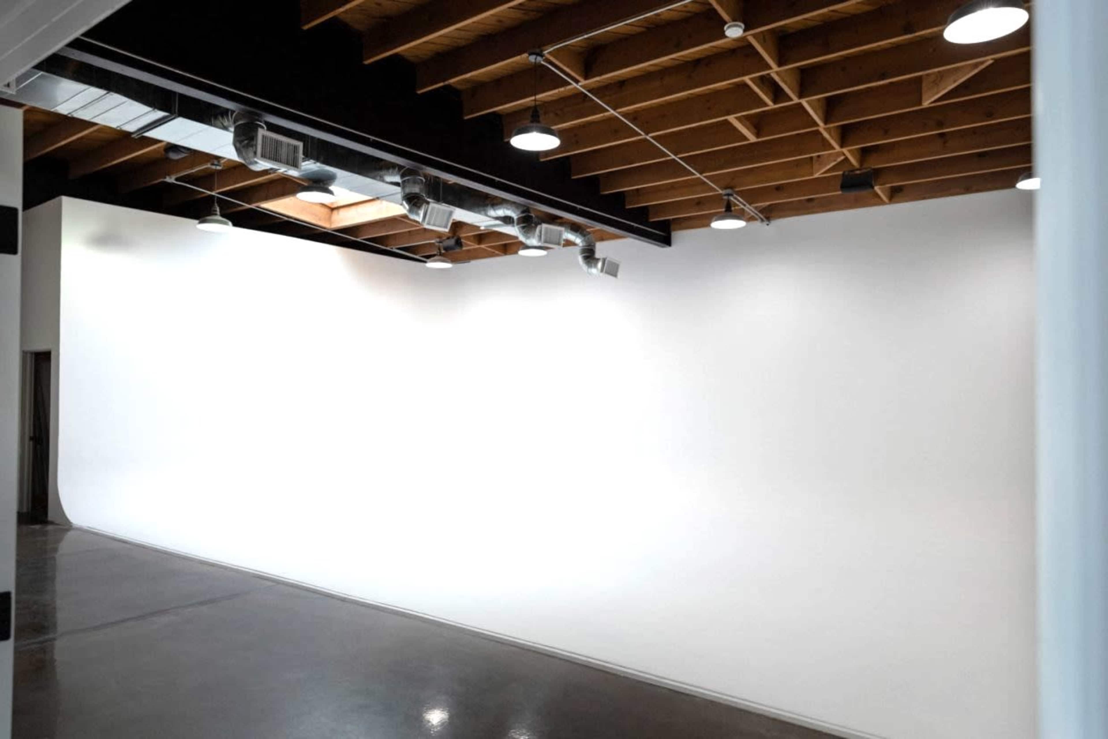 The image shows a spacious, indoor photography studio featuring a seamless white backdrop and wooden ceiling beams with track lighting.