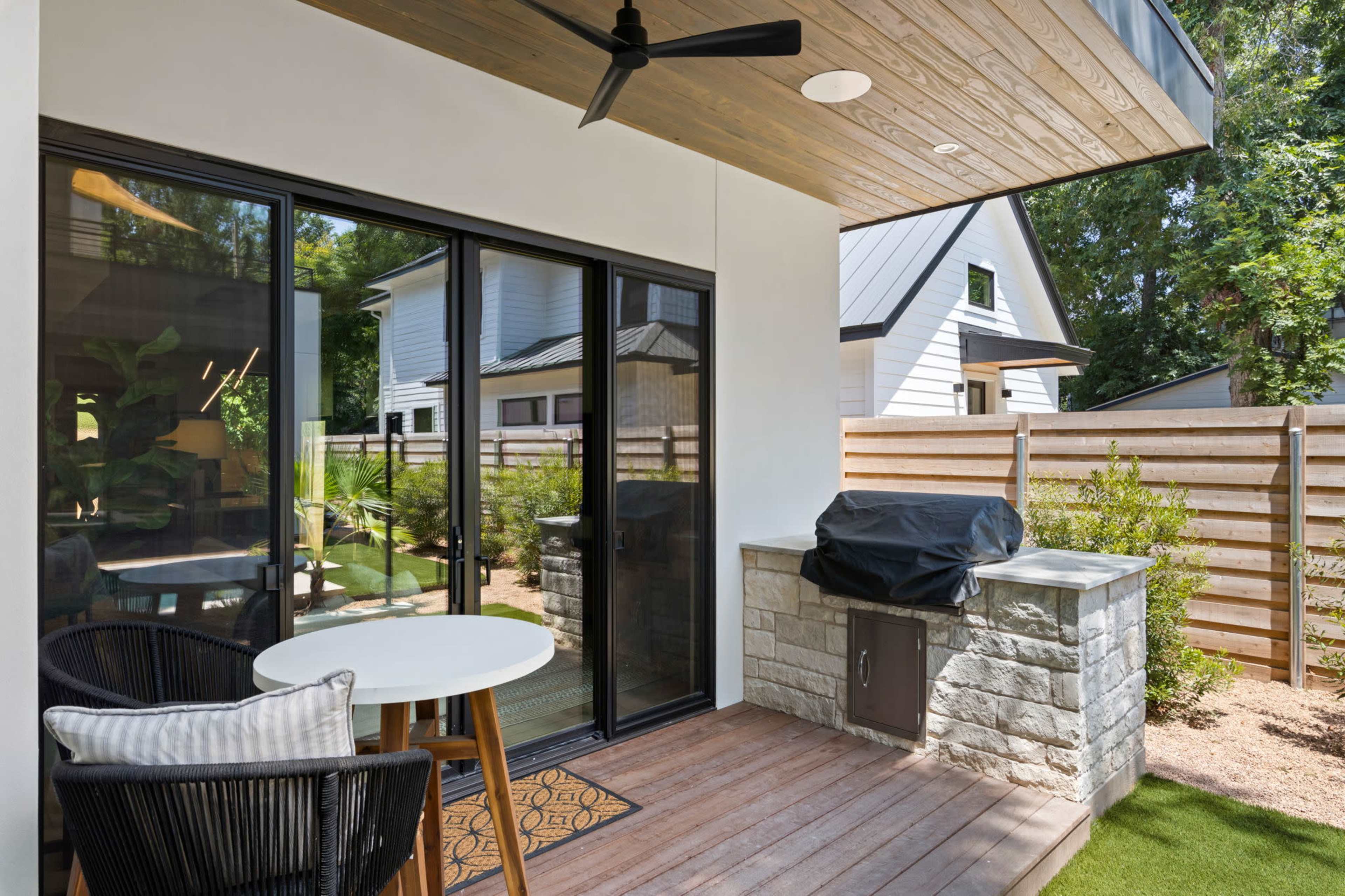 The image shows a patio area with a table and chairs, a built-in grill, and a view of a landscaped yard enclosed by a wooden fence.