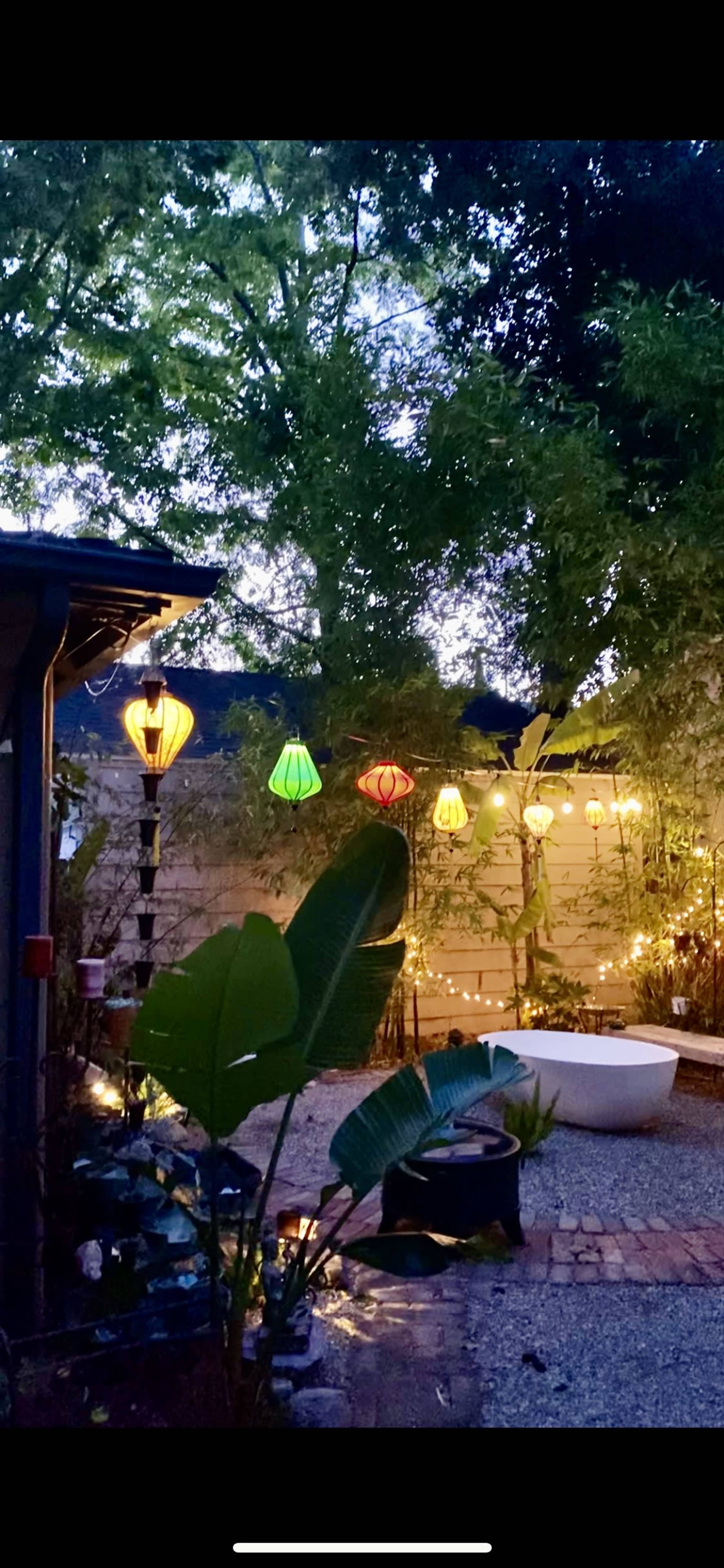 A tranquil backyard scene illuminated by colorful lanterns hanging among trees, with a white bathtub and a bench visible in the space.