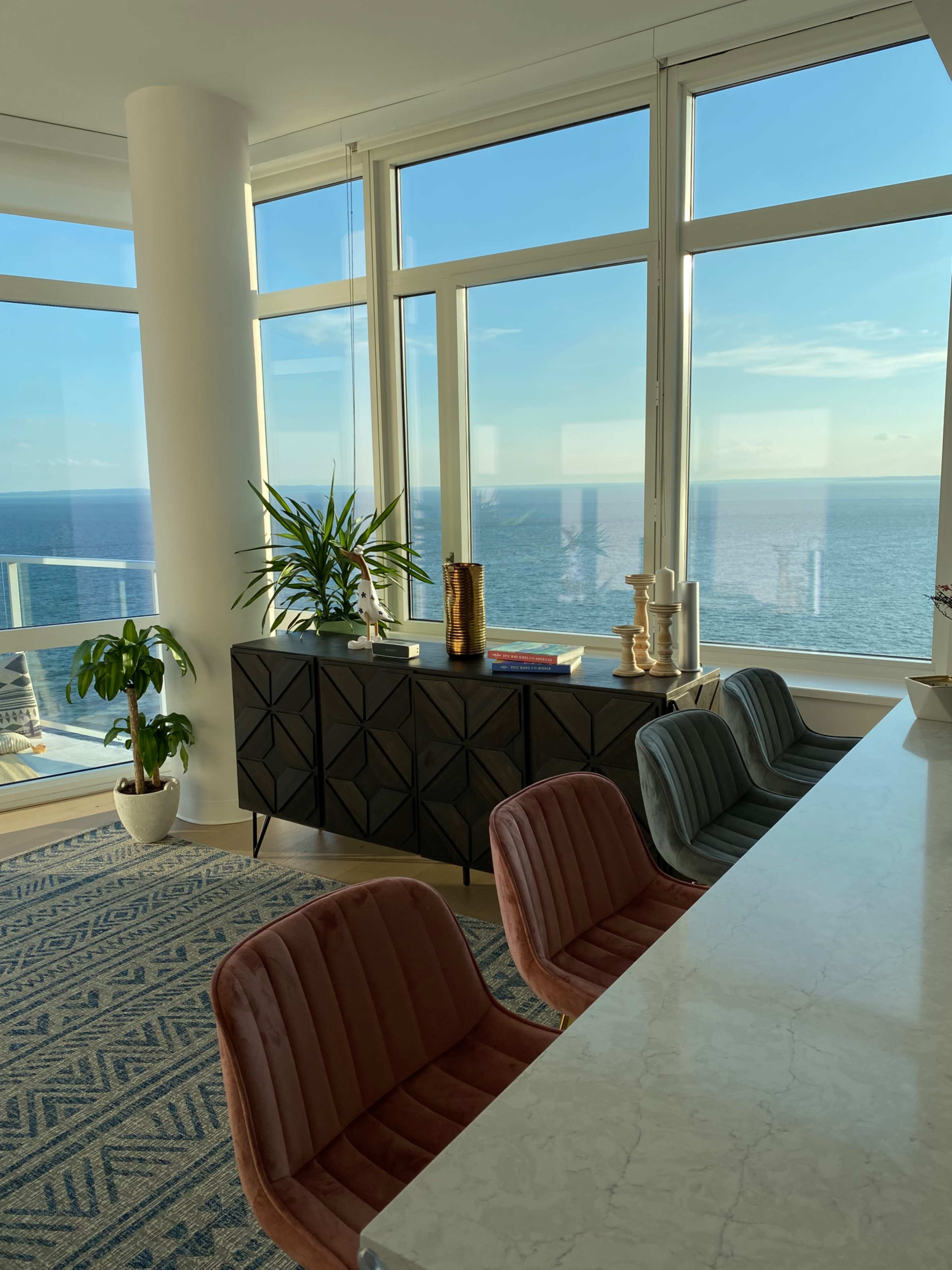 A modern interior with a view of the ocean, featuring a marble countertop, patterned rug, and stylish chairs near large windows.