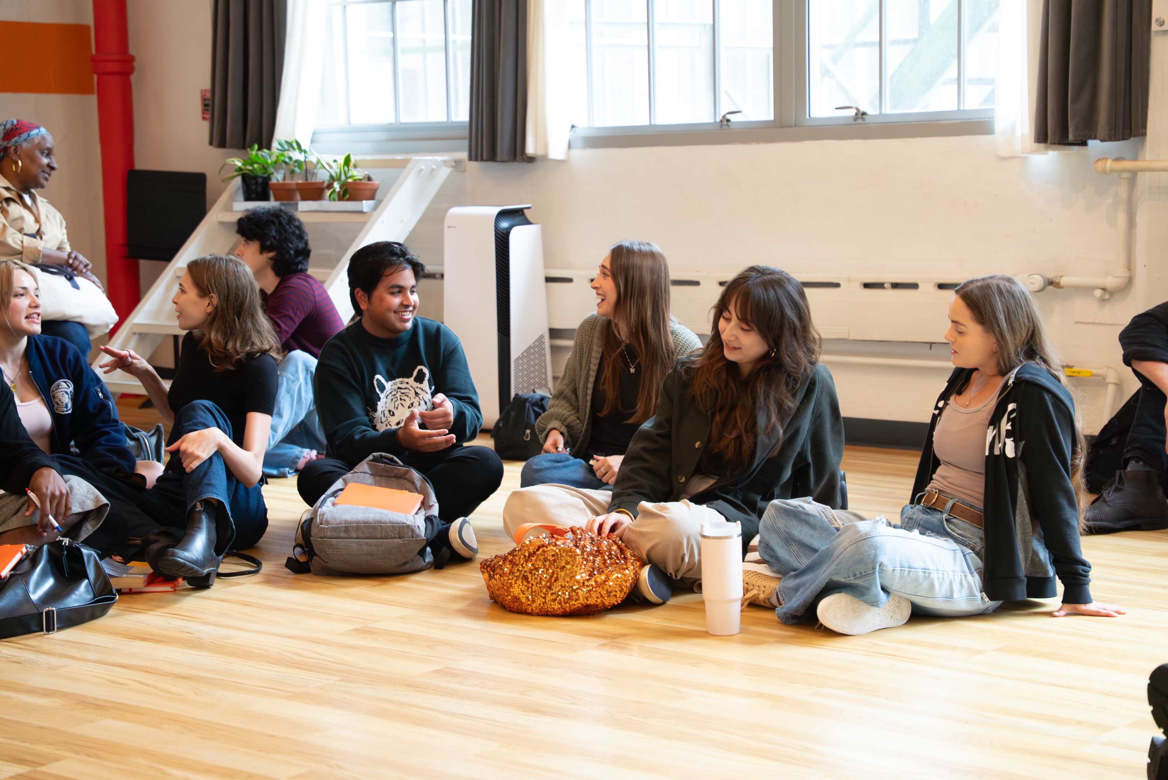 A group of young adults sits on the wooden floor in a bright room, engaging in conversation and laughter.