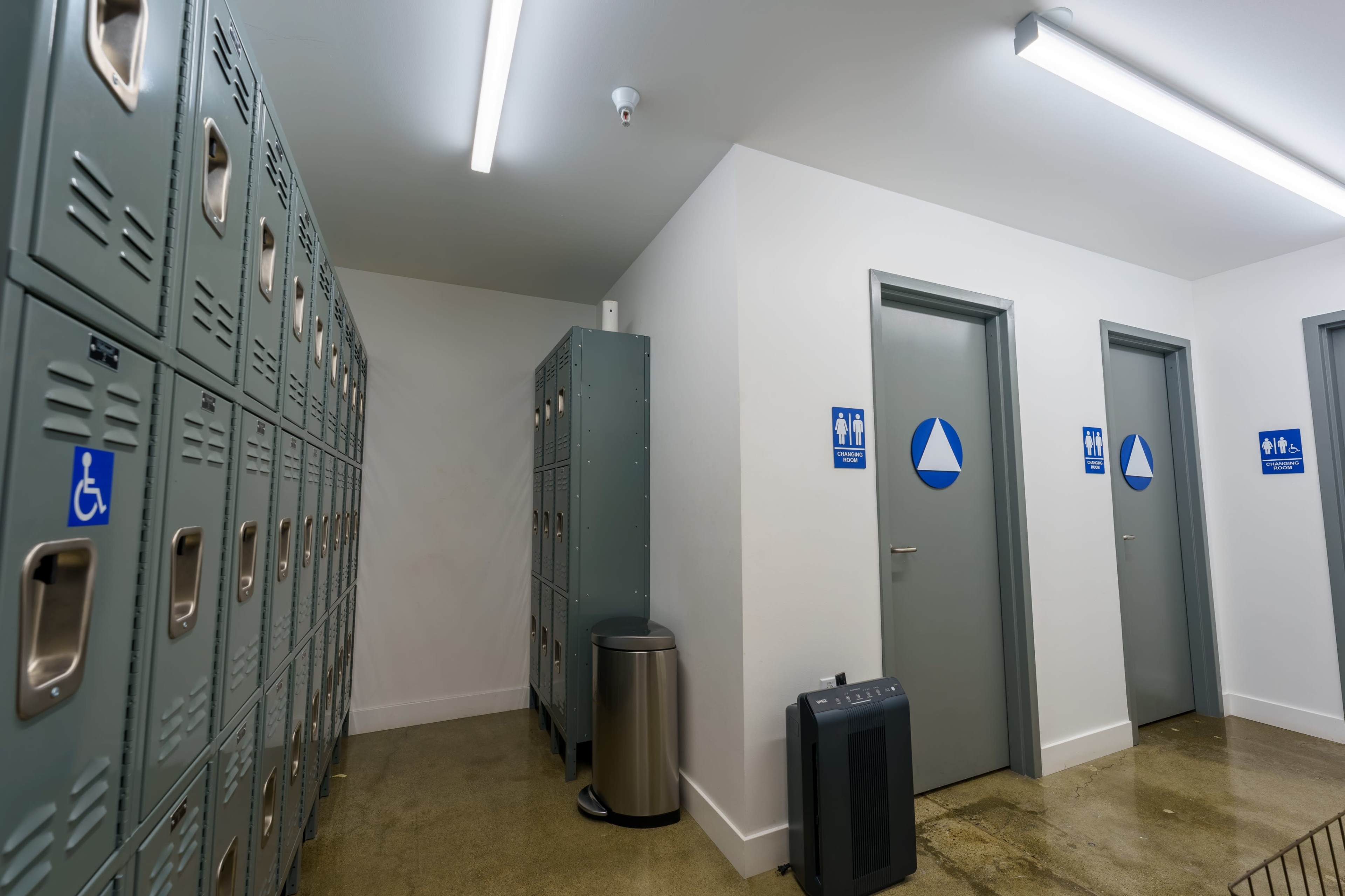 The image shows a hallway with metal lockers on the left, two restroom doors on the right, and a trash can nearby.