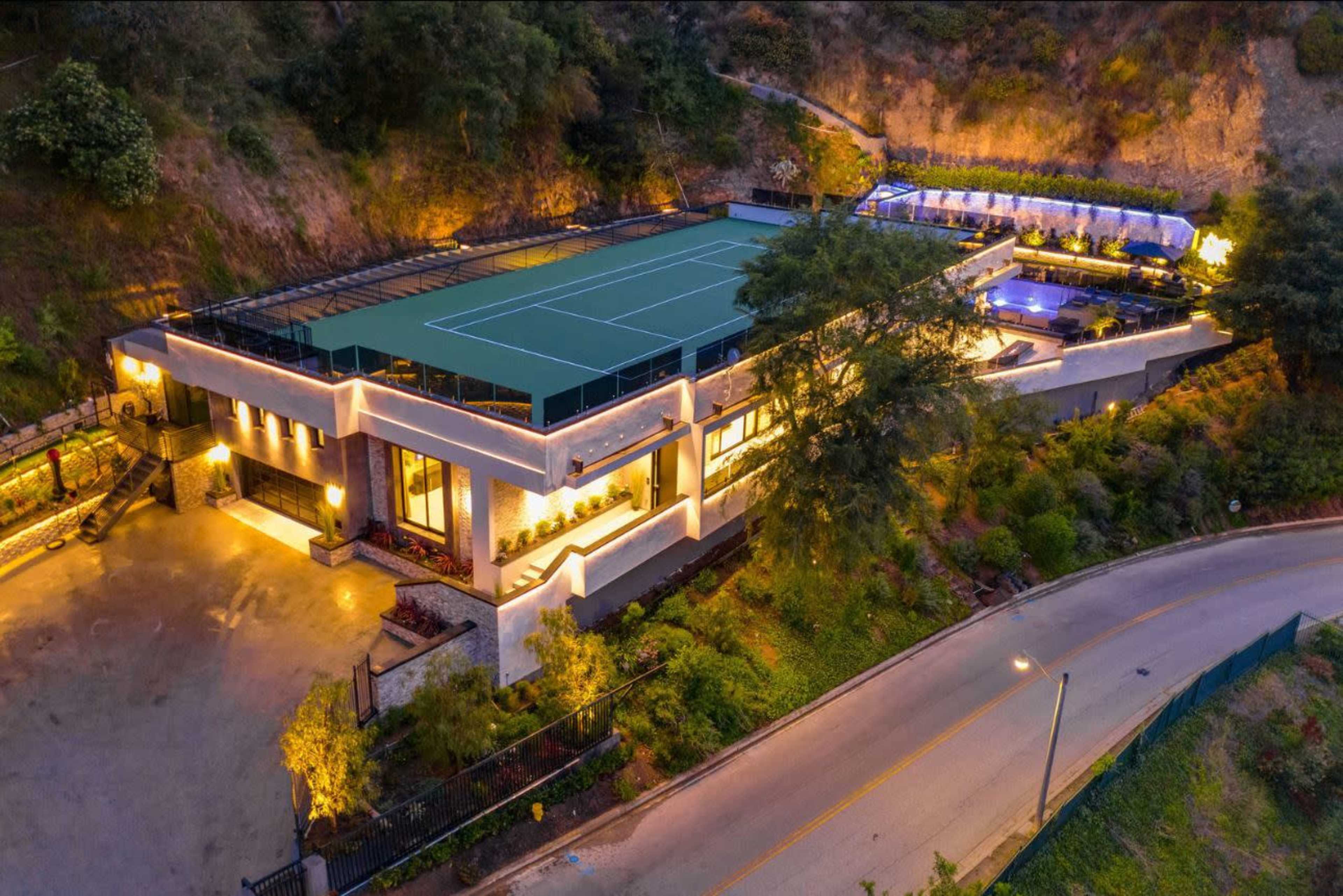 The image shows a large modern house with a rooftop tennis court, surrounded by landscaped gardens, and featuring a pool area illuminated at night.