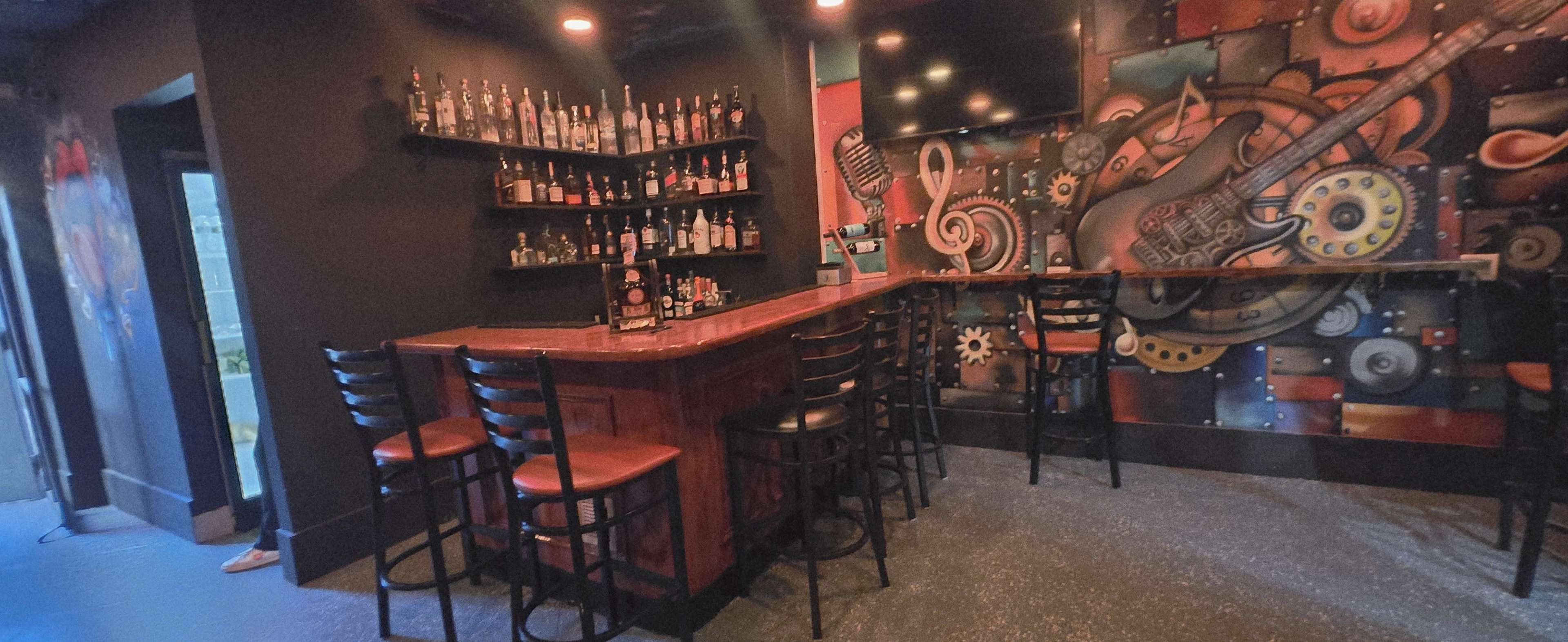 The image shows a dimly lit bar area with a wooden counter, a wall adorned with vibrant artistic murals, and several black bar stools lined up in front.