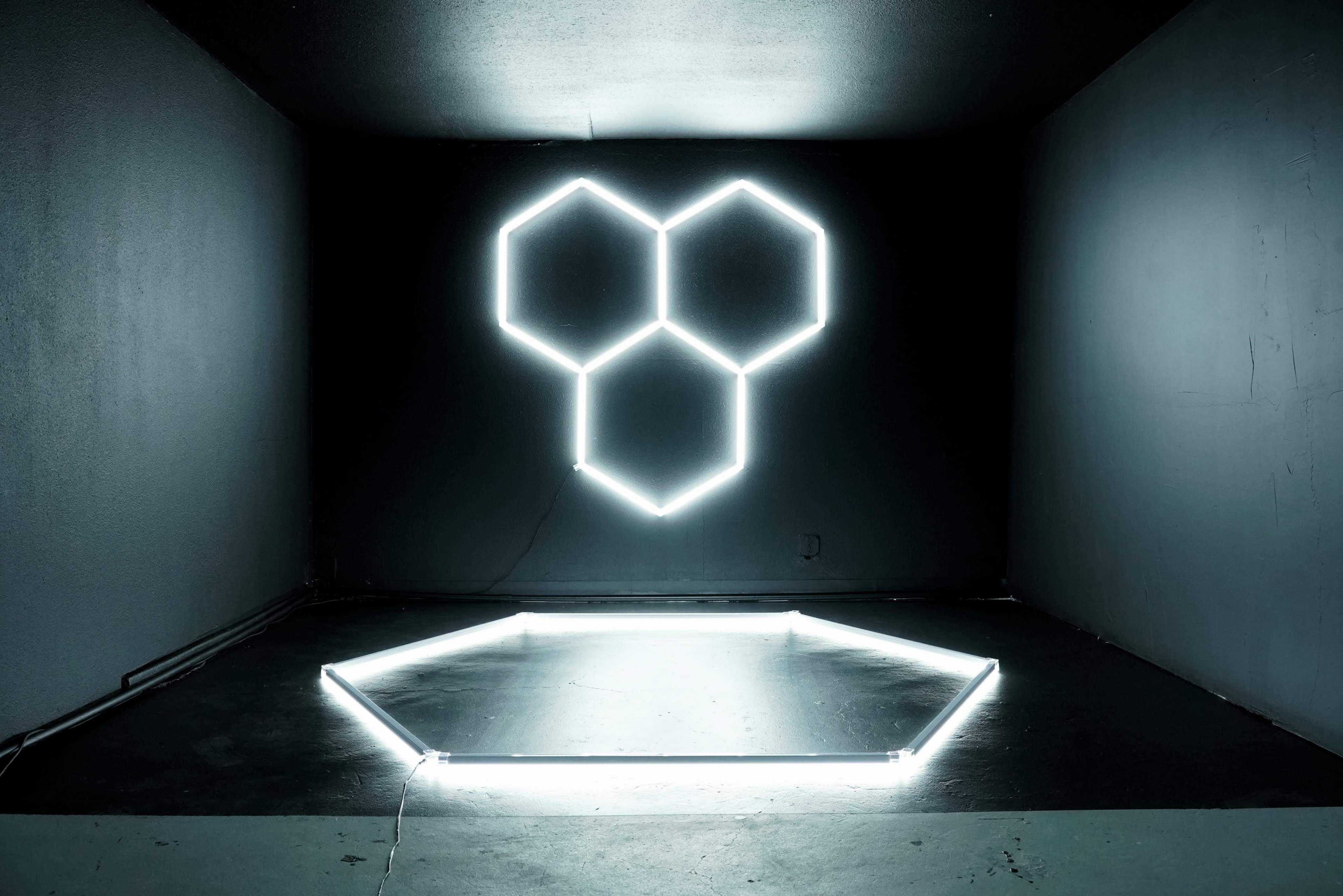 The image shows a dark room with a hexagonal light installation on the wall and a corresponding hexagonal light outline on the floor.