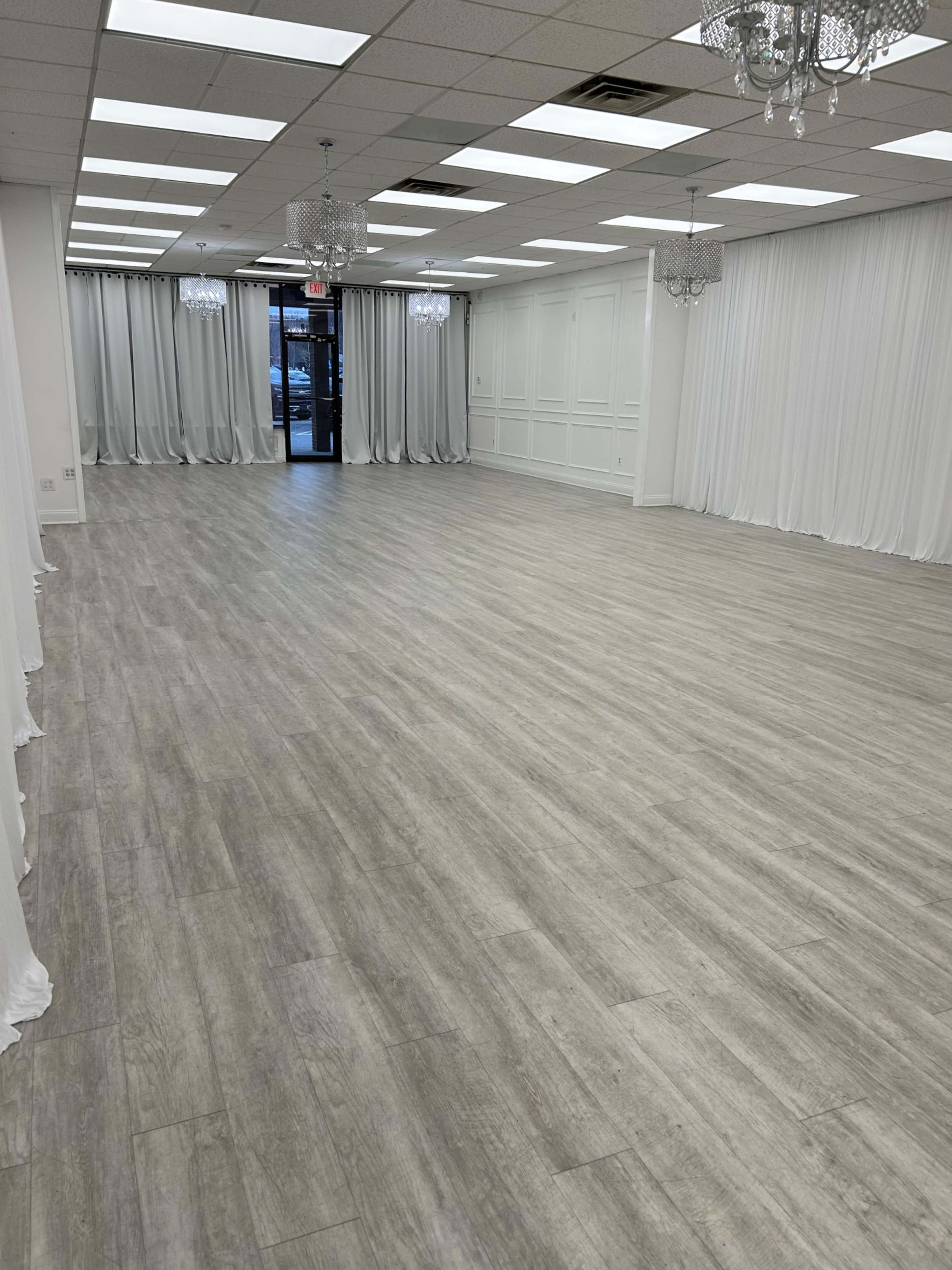 The image shows a spacious, empty interior room with wooden flooring, white drapes along the walls, and several hanging chandeliers.