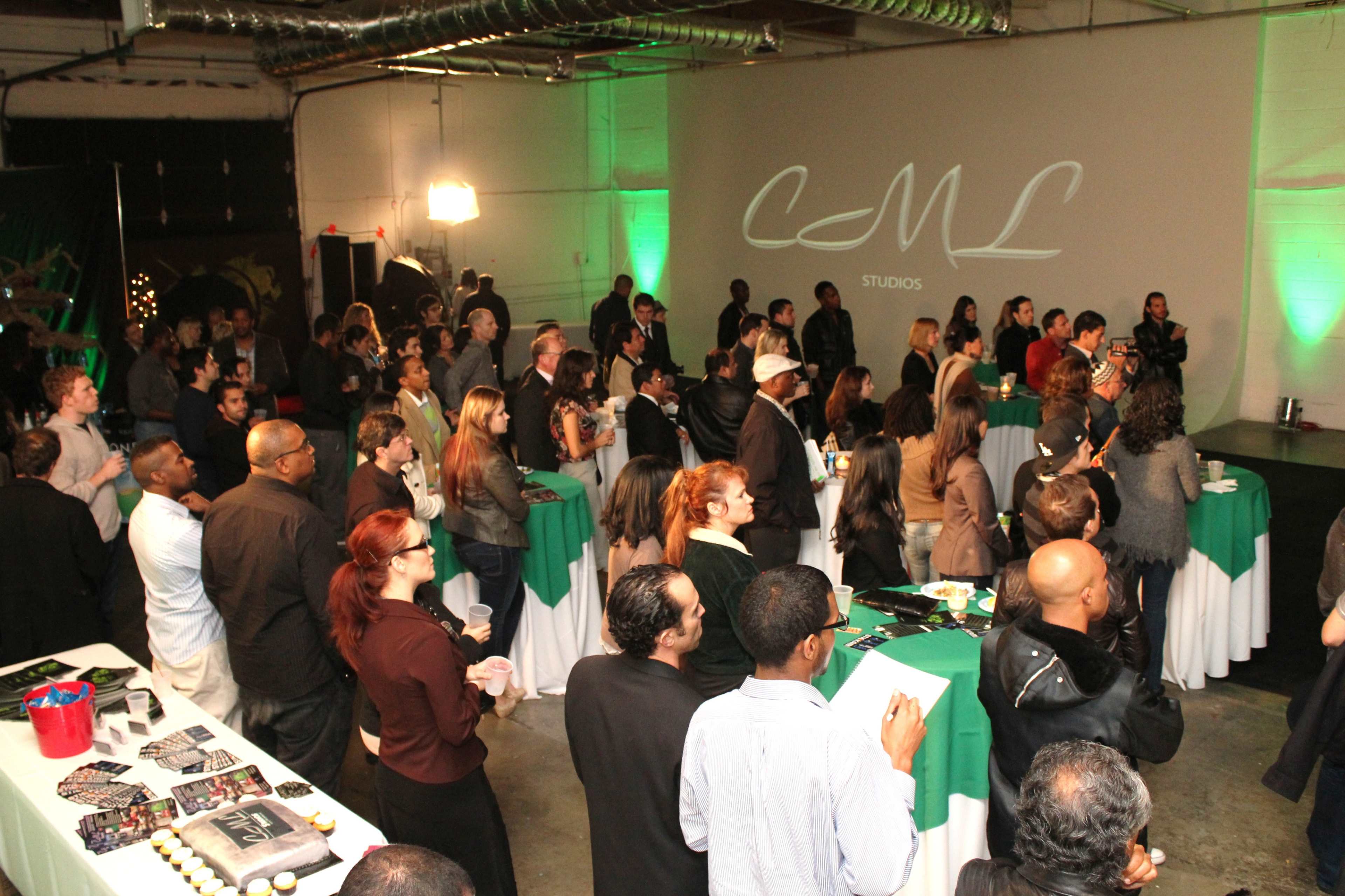A large group of attendees stands in a studio space, facing a presentation screen in an event setting with tables covered in green and white.