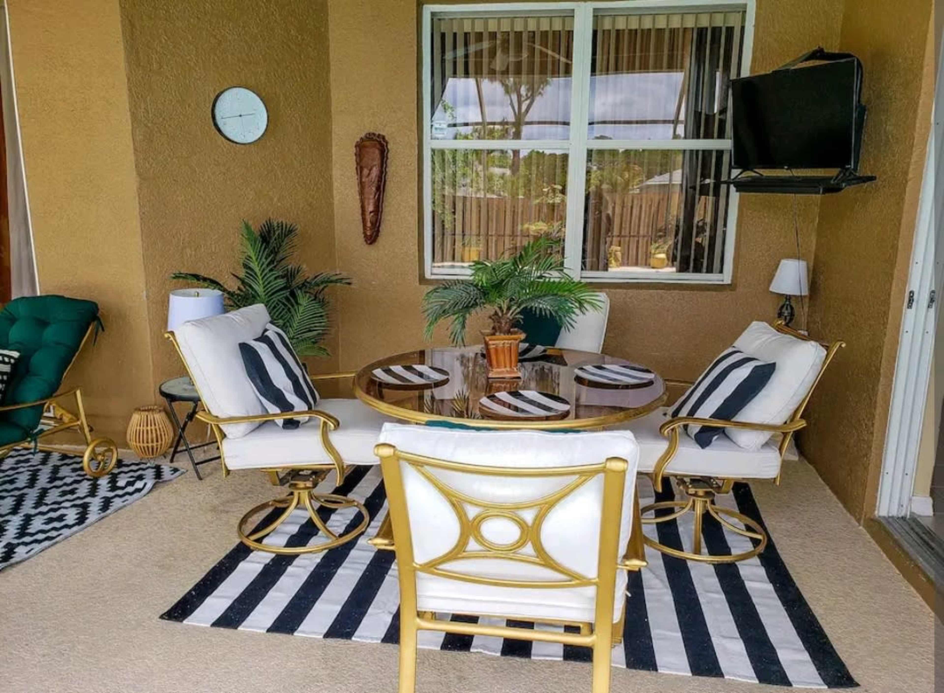 A cozy outdoor patio featuring a round glass table surrounded by white and black chairs, with a patterned rug underneath and several decorative plants.