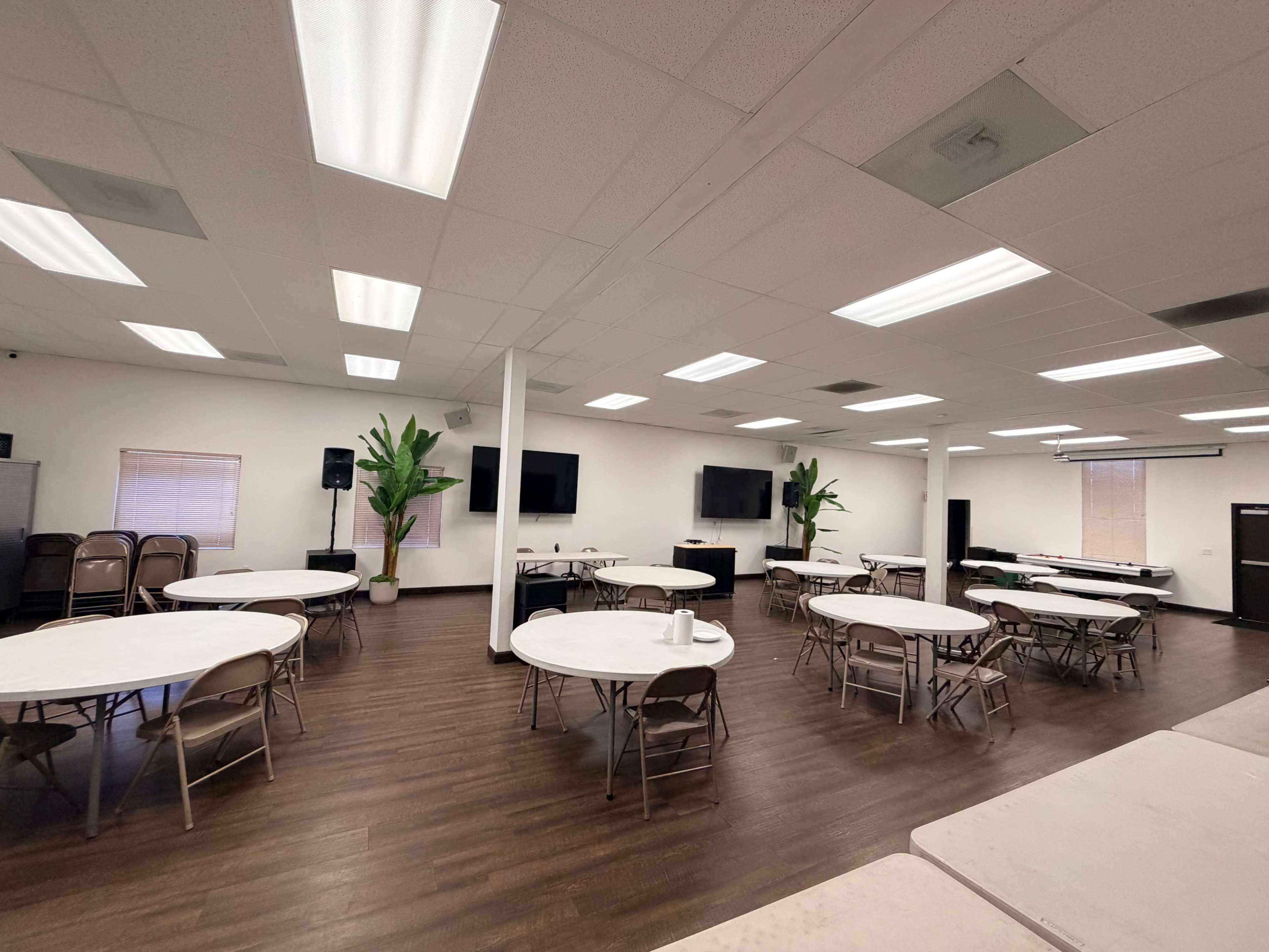 The image shows a spacious room with several round tables surrounded by chairs, equipped with two large screens on the walls and a potted plant in the corner.