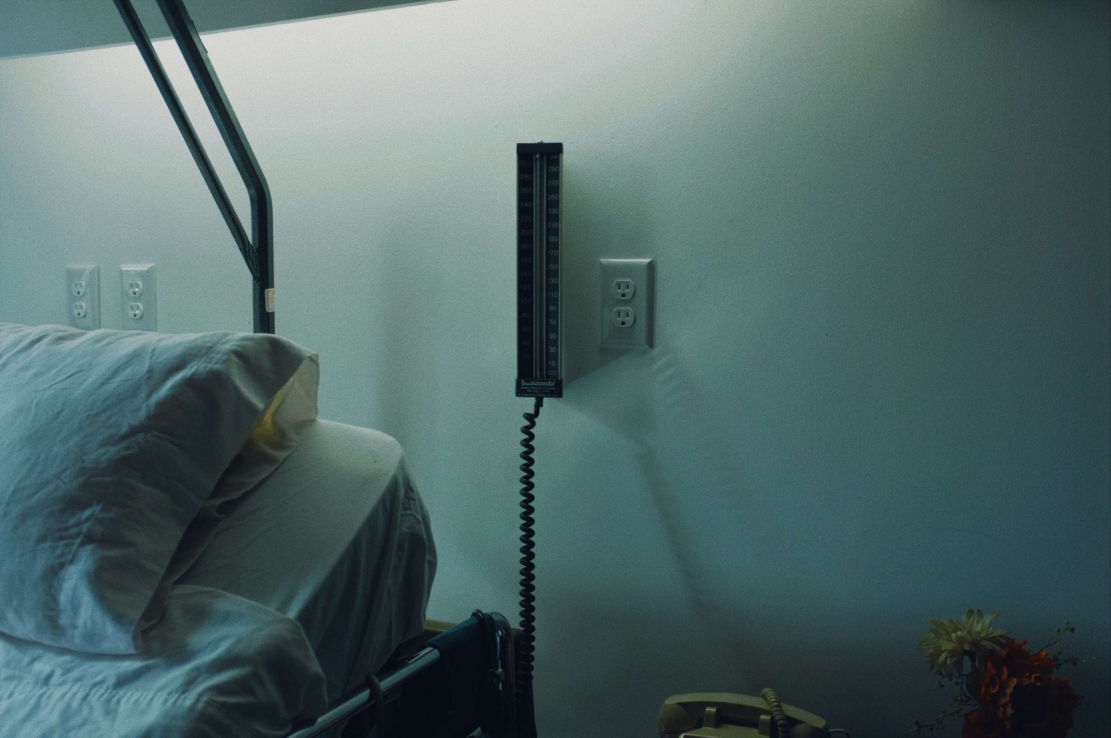 The image shows a hospital room with a bed and a wall-mounted sphygmomanometer beside a power outlet.