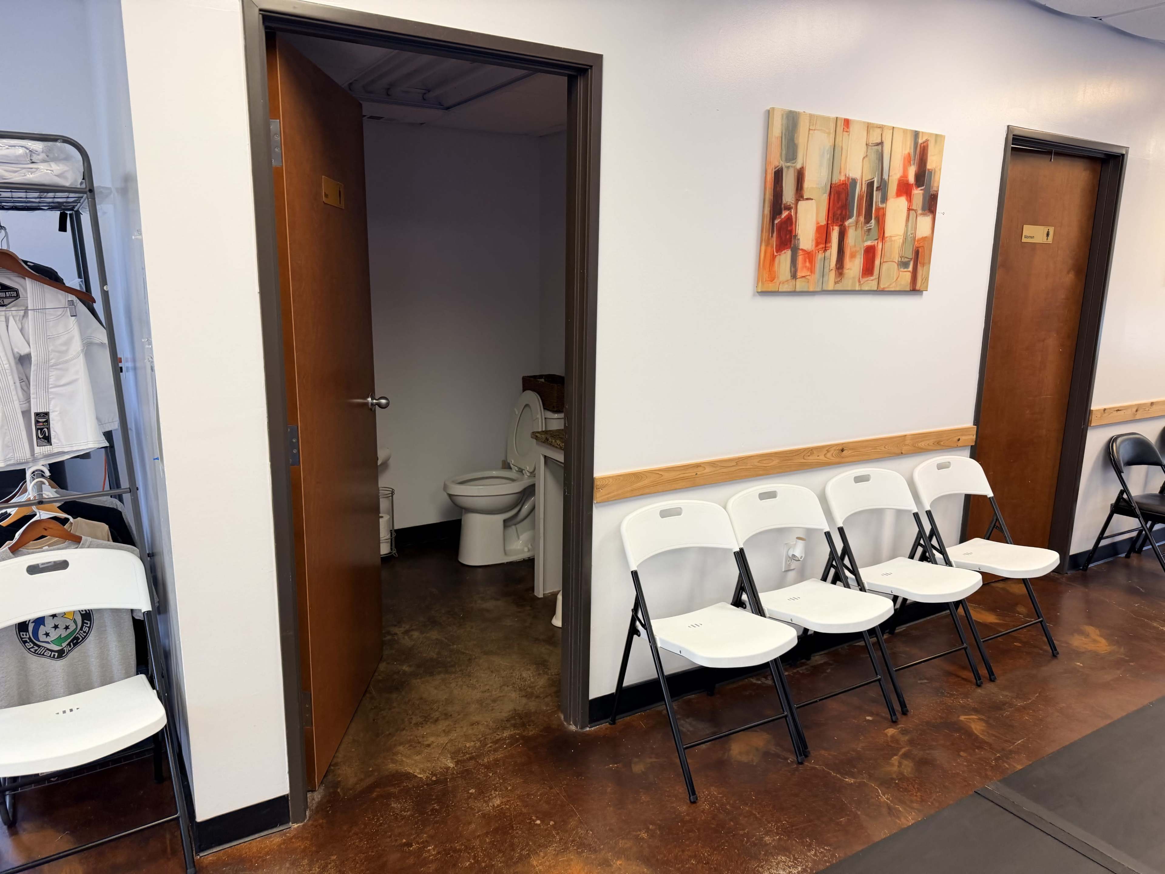 The image shows a waiting area with several folding chairs facing a restroom door and another door, along with a wall-mounted artwork.