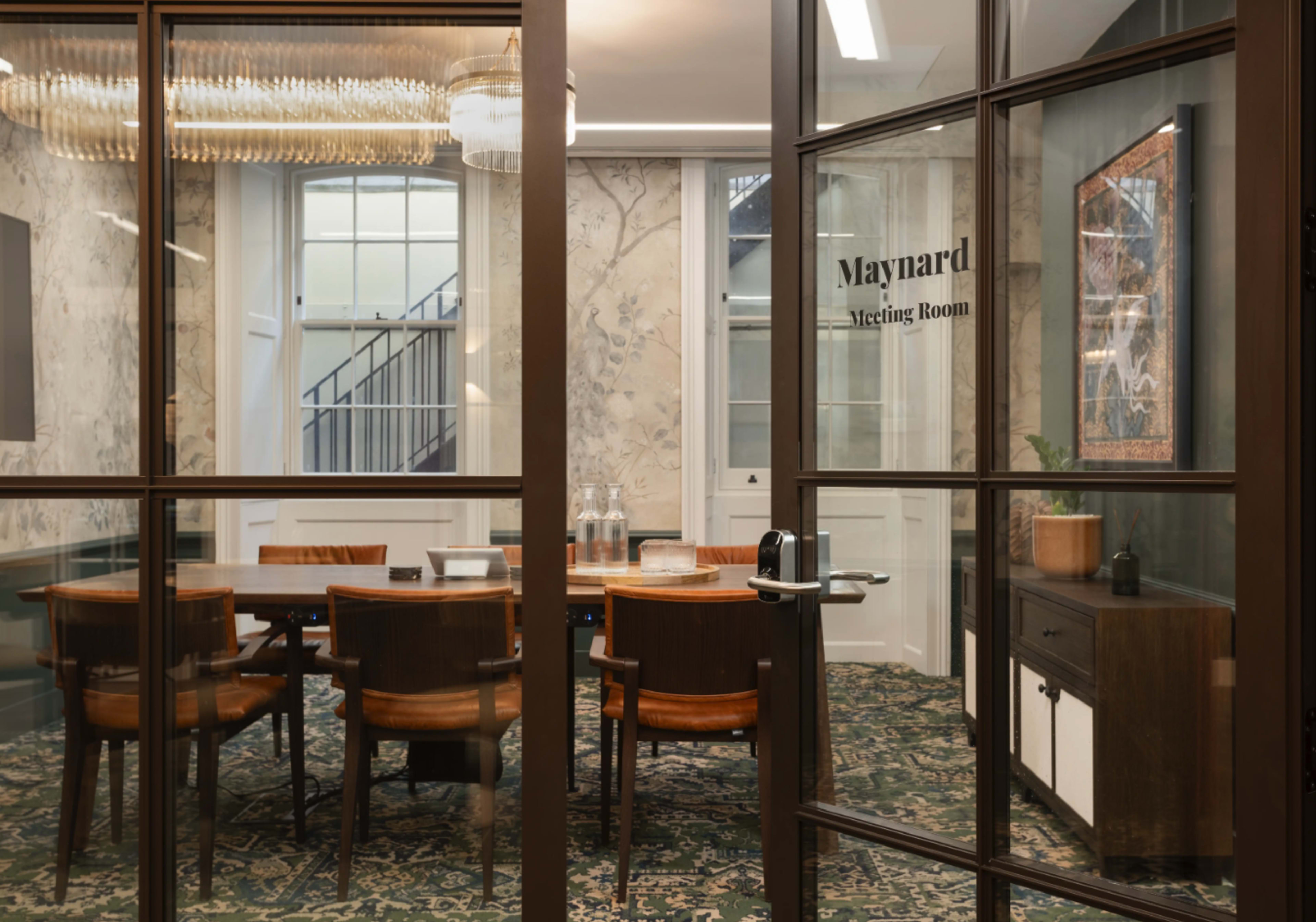 The image shows a modern meeting room with a large wooden table surrounded by chairs, located behind glass doors featuring a nameplate.
