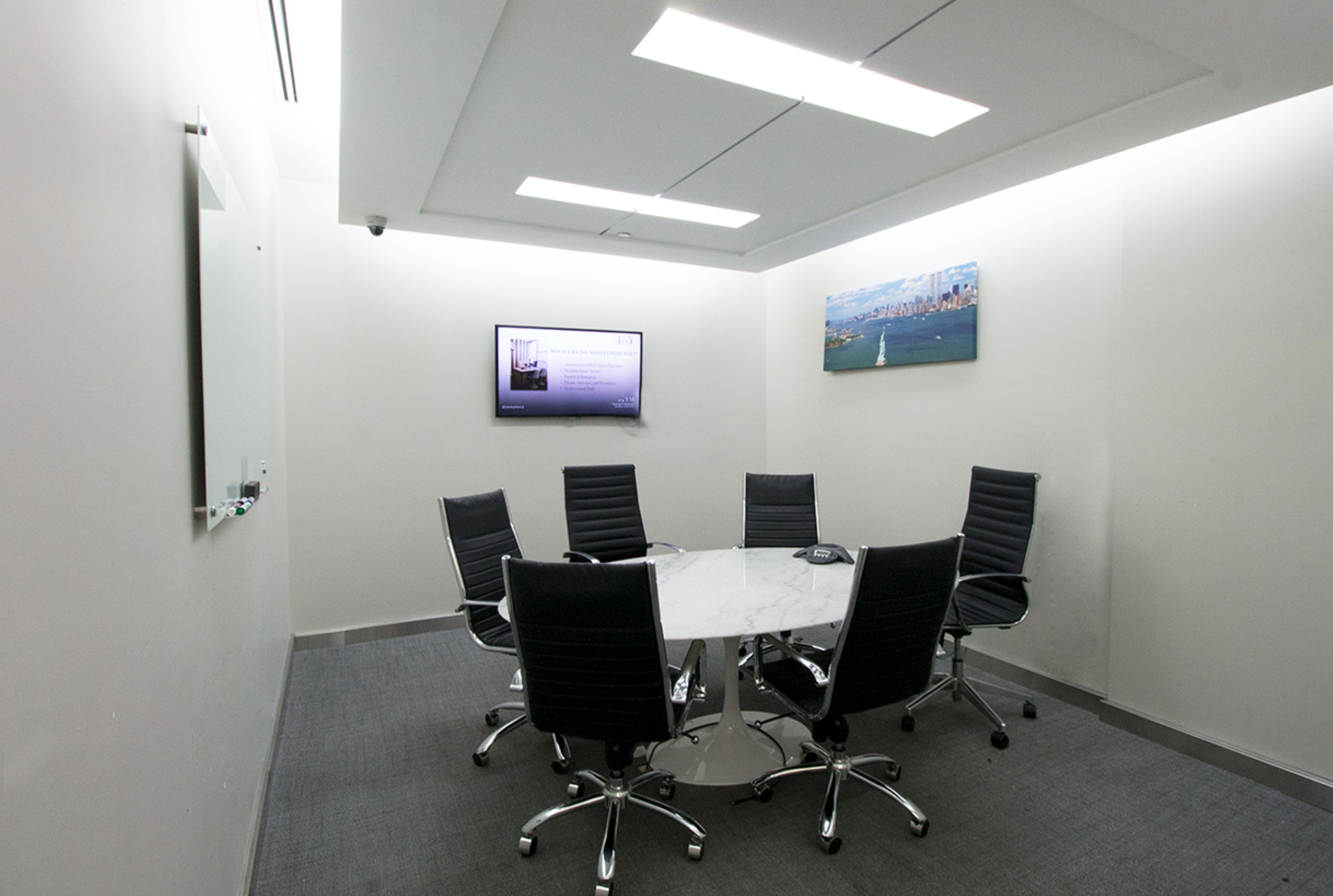 A small conference room features a round marble table surrounded by six black swivel chairs, with a monitor and wall art displayed on the walls.