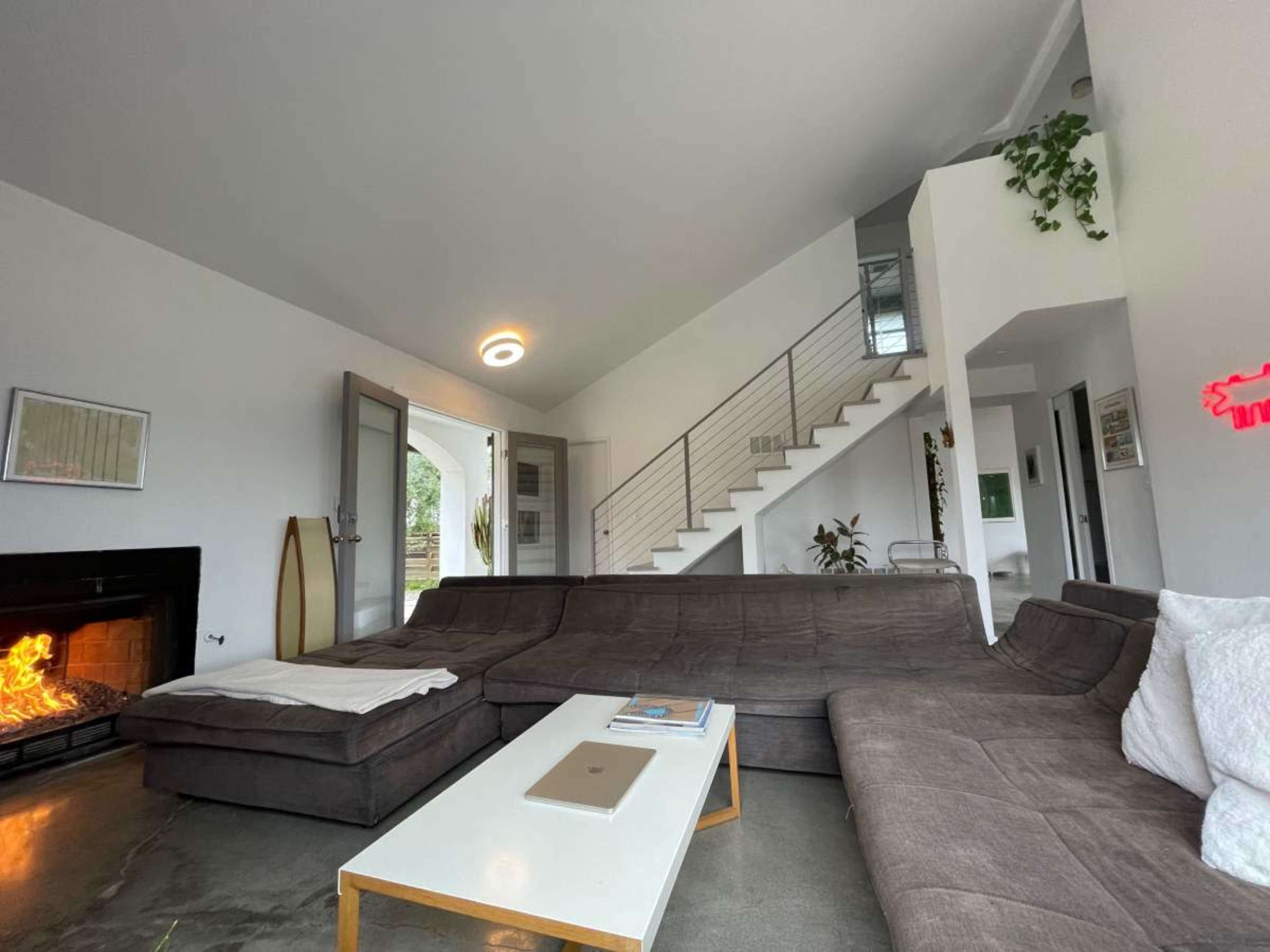 The image shows a modern living room featuring a large sectional sofa, a coffee table, a fireplace, and a staircase leading to an upper level.