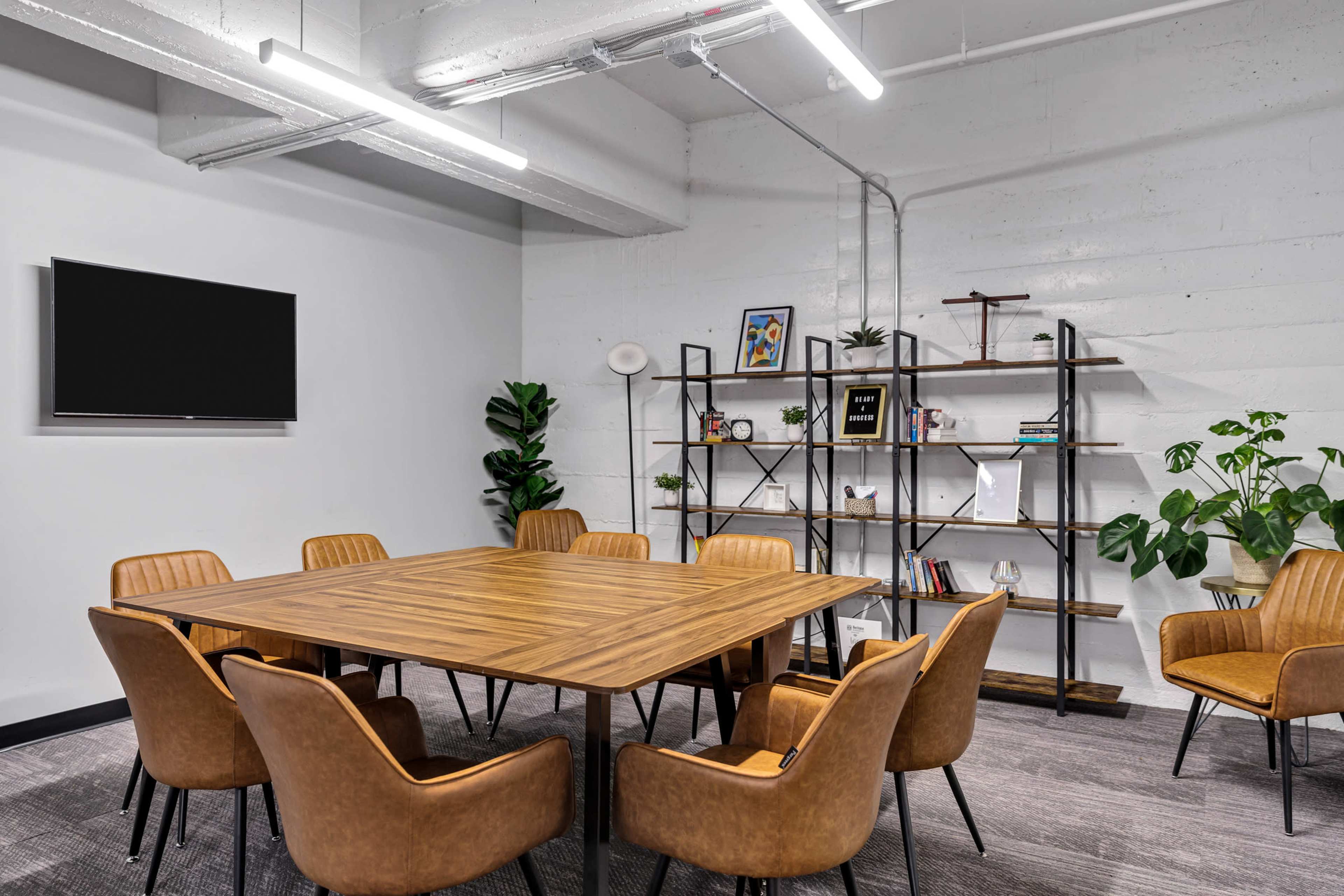 The image shows a modern conference room with a large wooden table surrounded by brown chairs, a wall-mounted TV, and a shelving unit filled with books and decorative items.