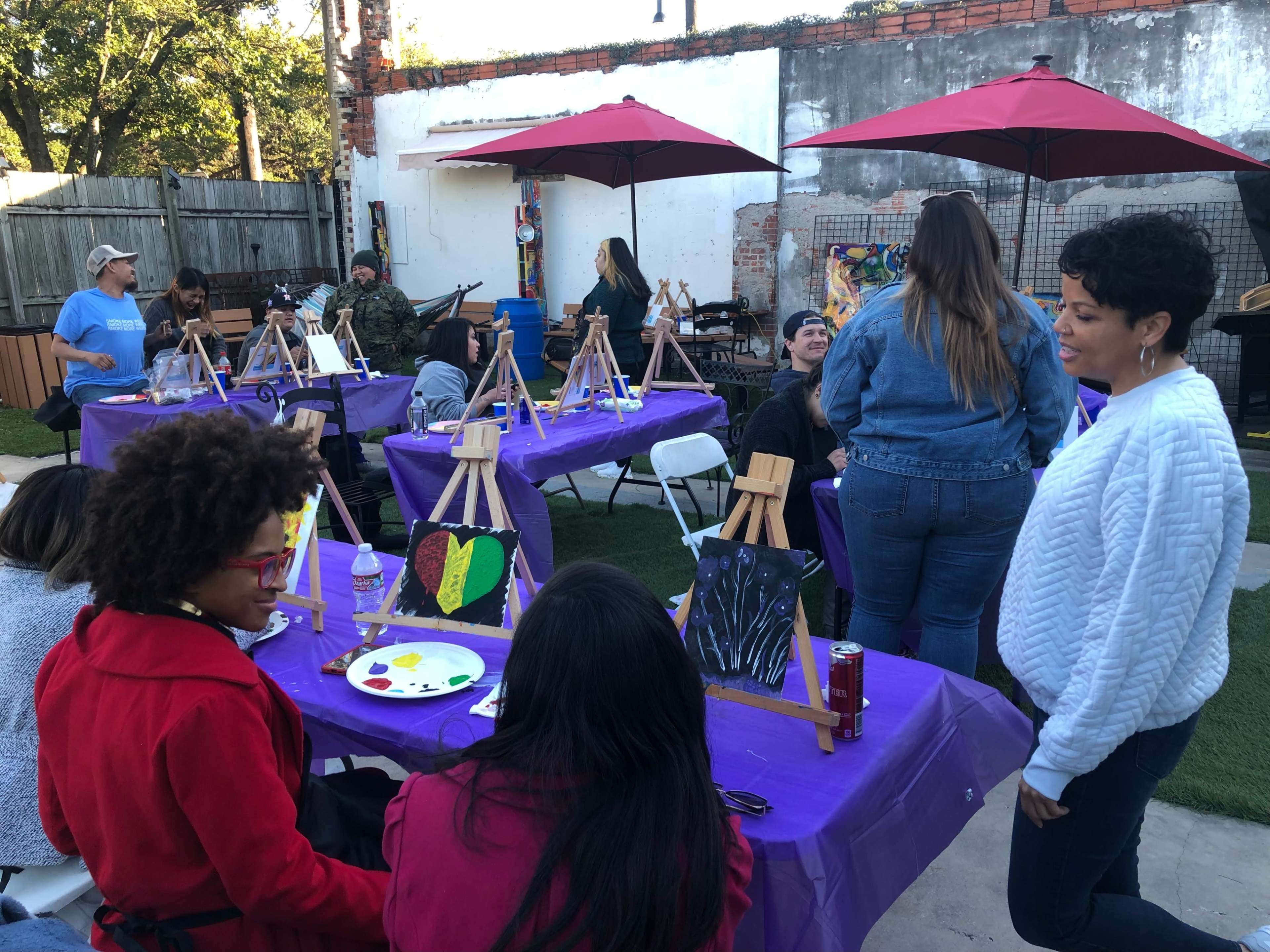 A group of people is engaged in an art class, with easels set up on tables covered in purple cloths and a variety of paintings displayed.
