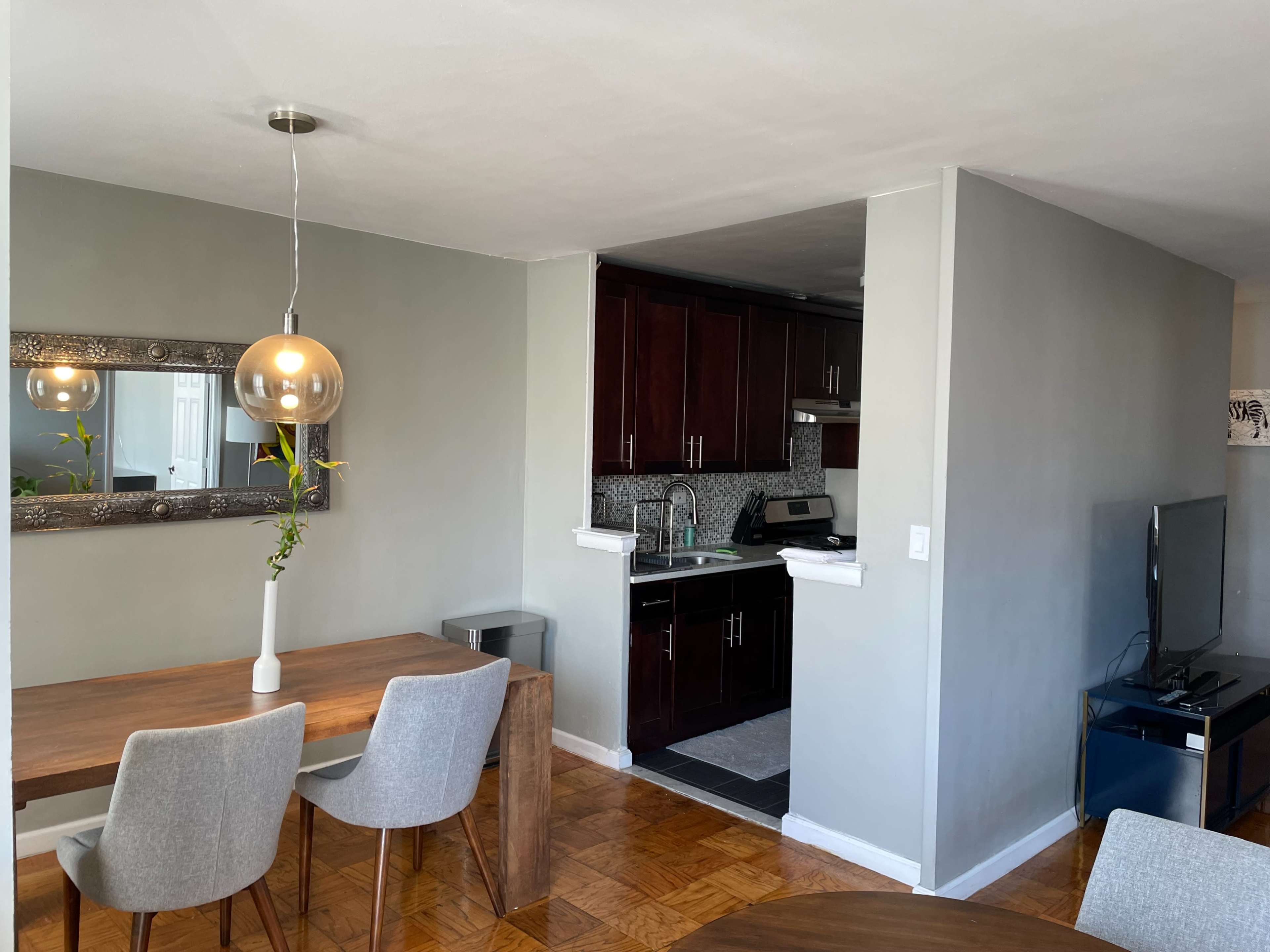 The image shows a modern dining area with a wooden table and gray chairs, adjacent to a small kitchen featuring dark cabinets and a countertop.