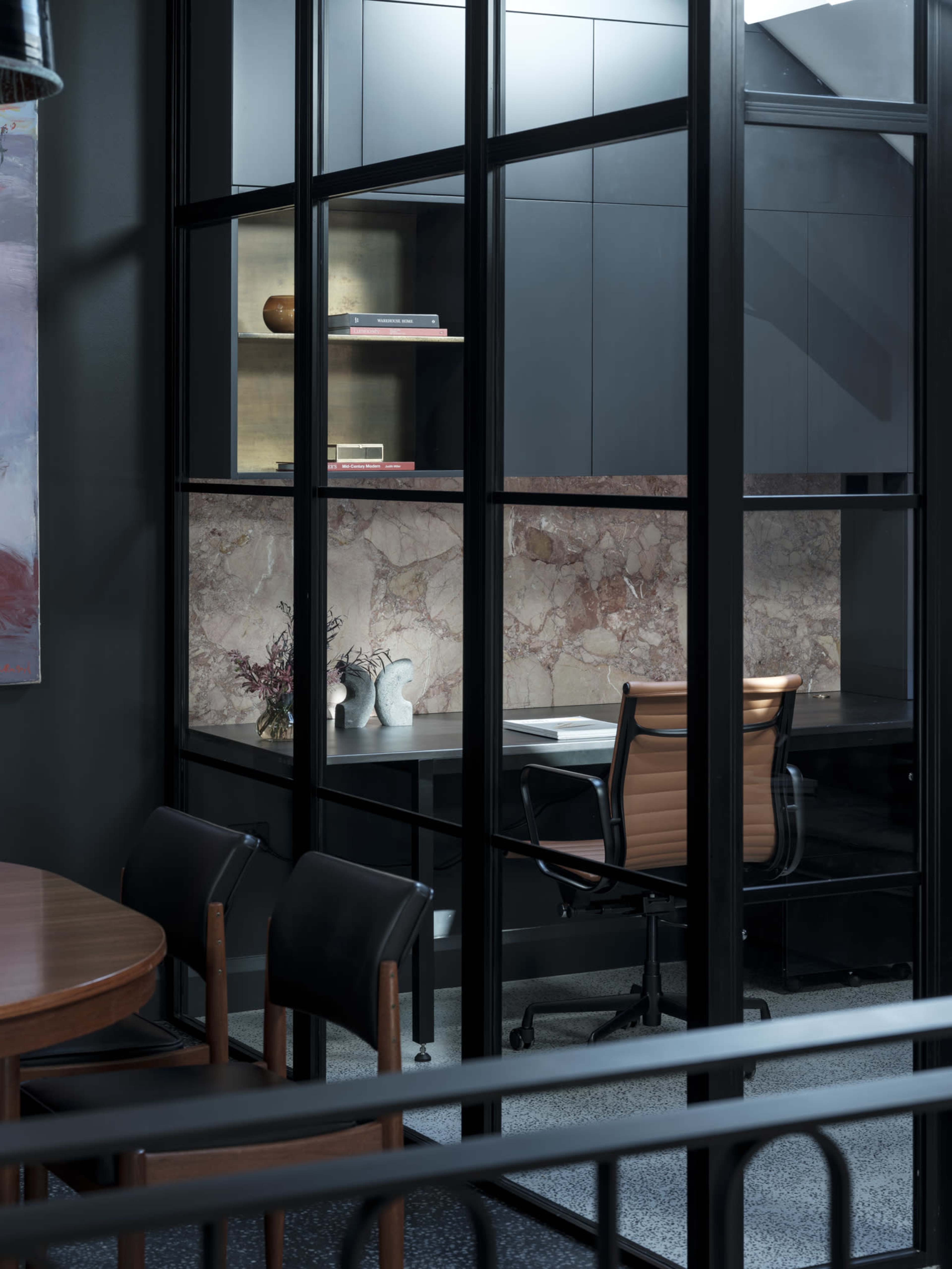 A modern office space features a glass-walled enclosure with a desk, an ergonomic chair, and a decorative marble wall, alongside a wooden dining table and chairs.