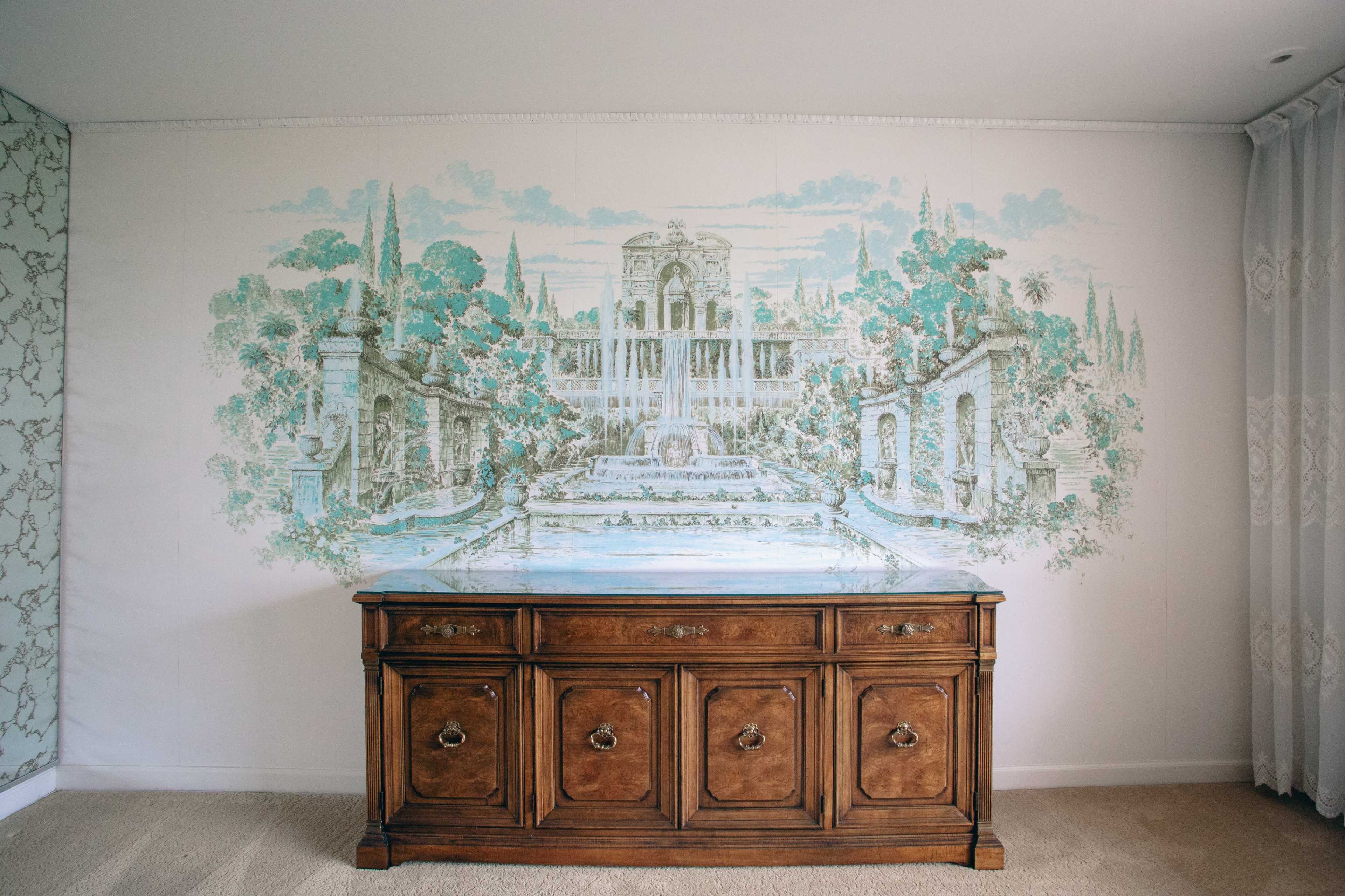 A wooden cabinet stands in front of a painted mural depicting an ornate garden with fountains and classical architecture.