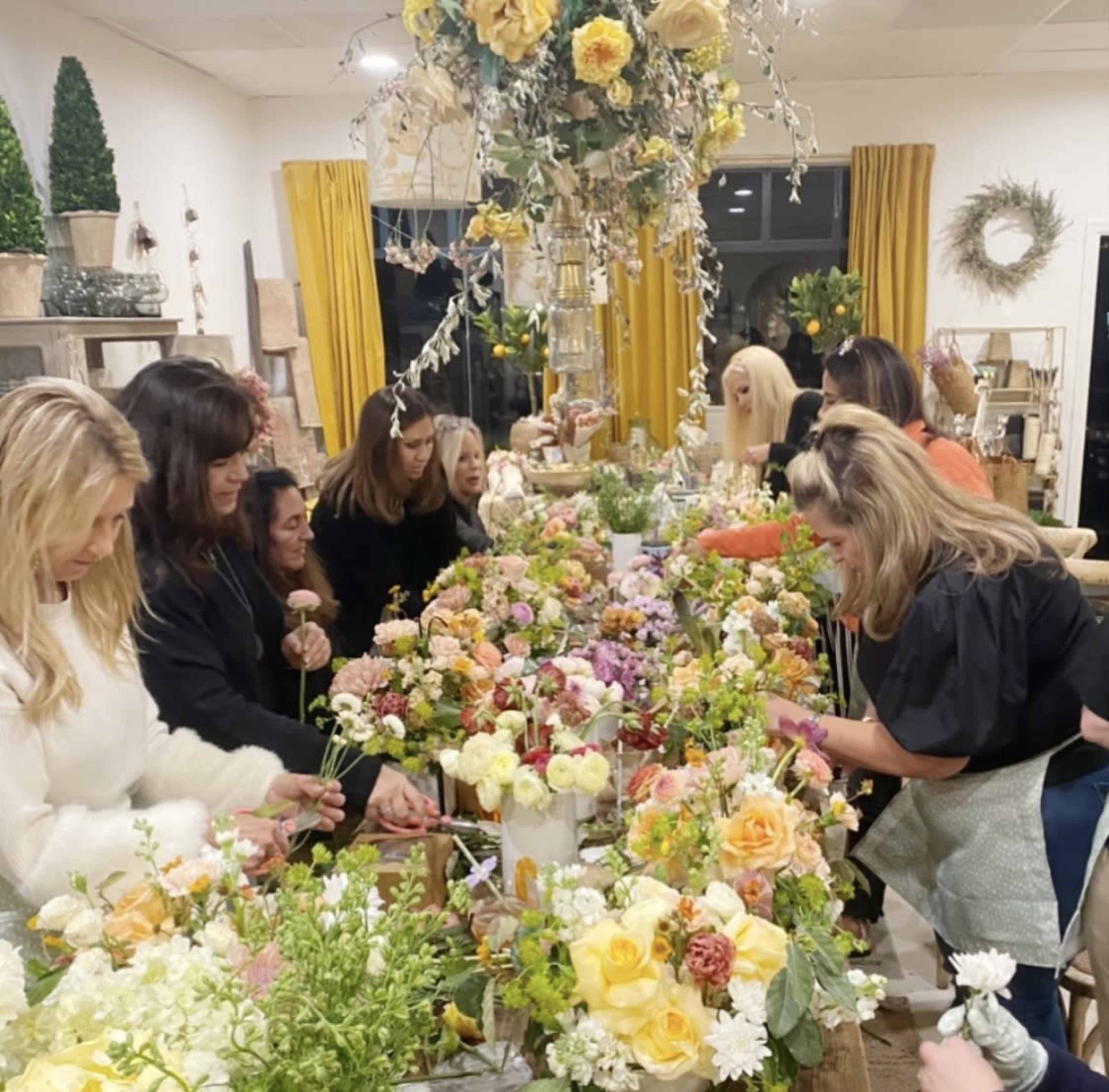 A group of women arranges flowers on a long table in a well-decorated room with bright yellow curtains.