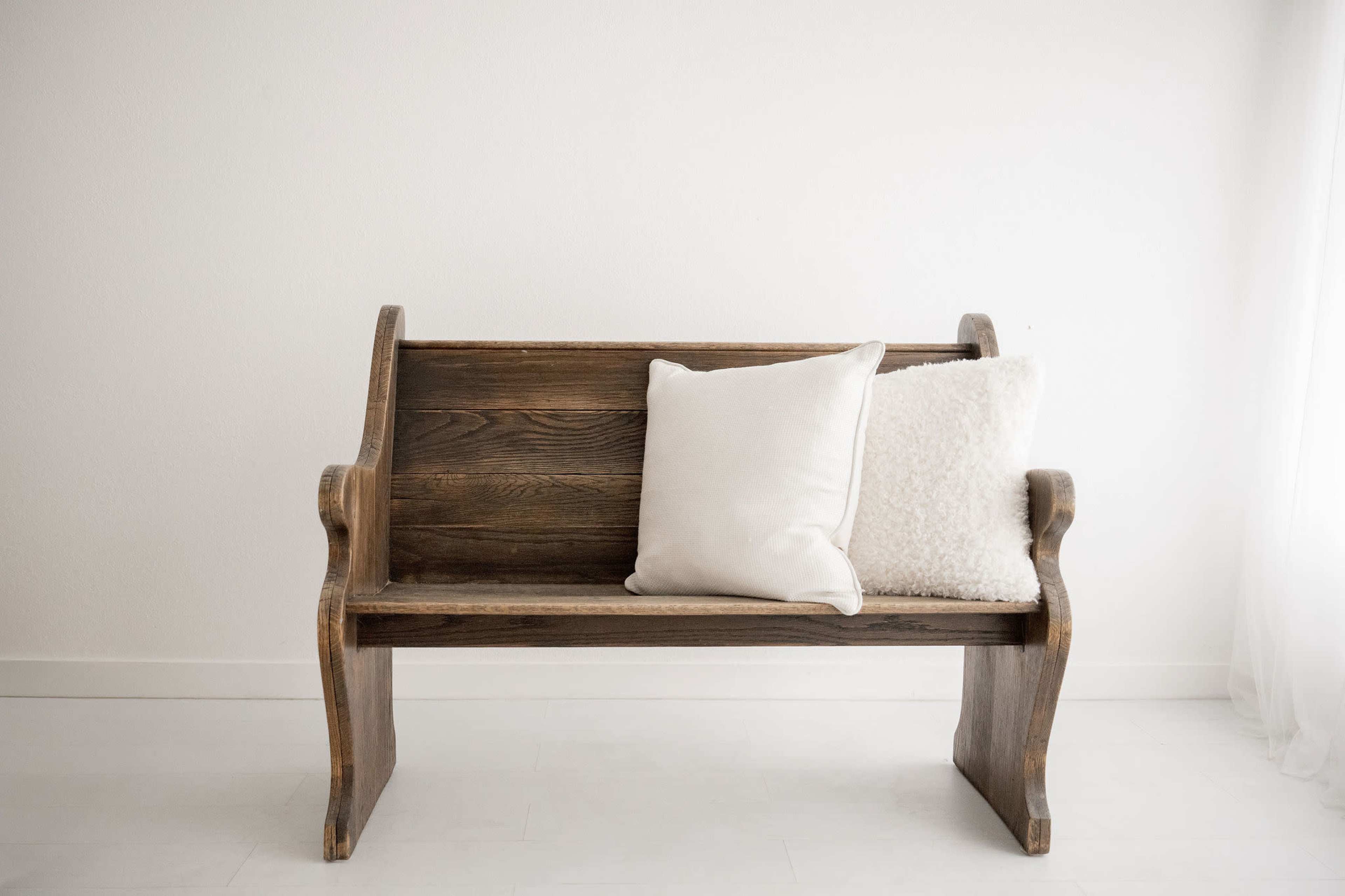 A wooden bench with a light finish and two pillows is positioned against a white wall.