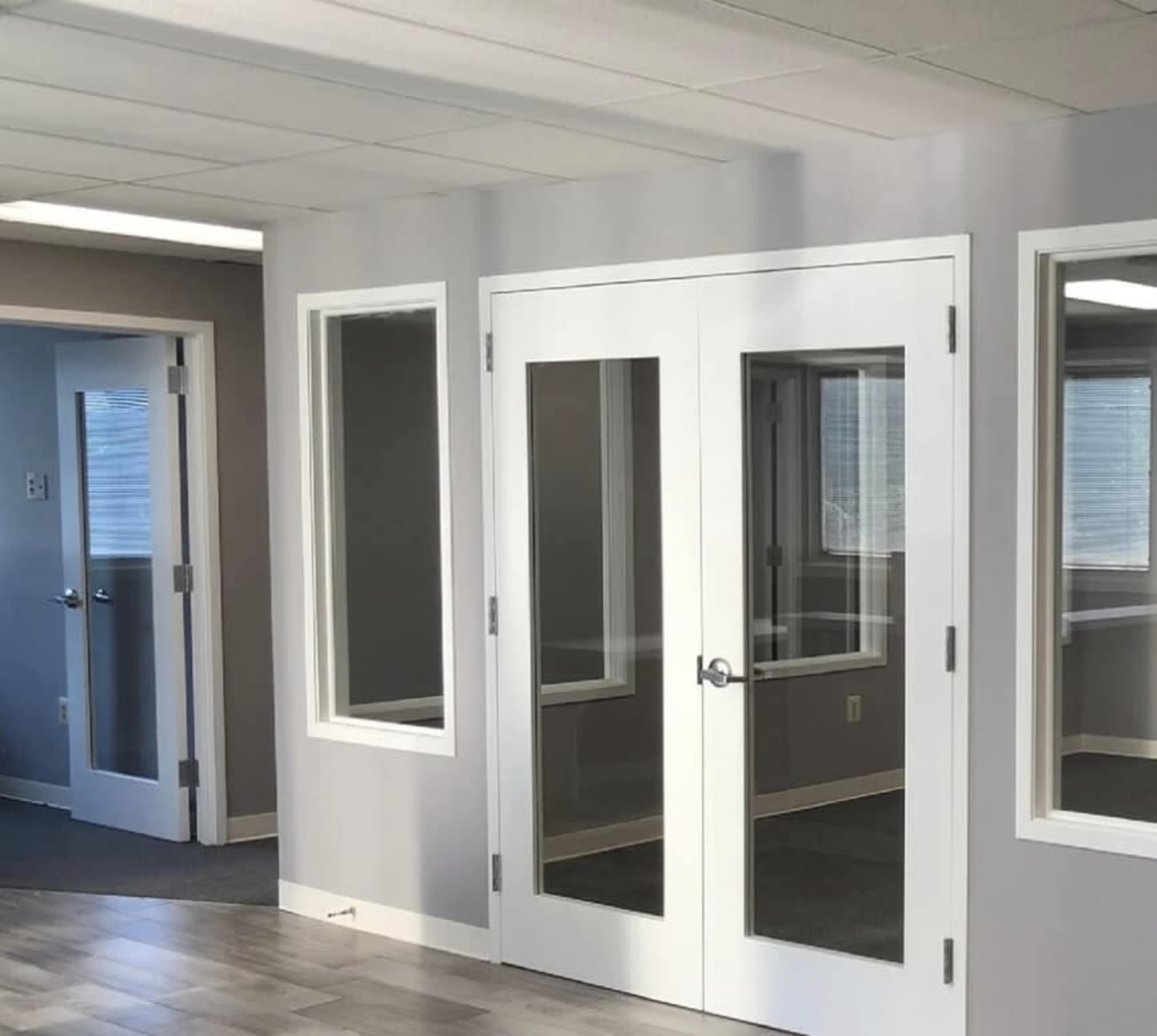 A modern office space featuring double glass doors and large windows with a light-colored wall and gray carpeting.