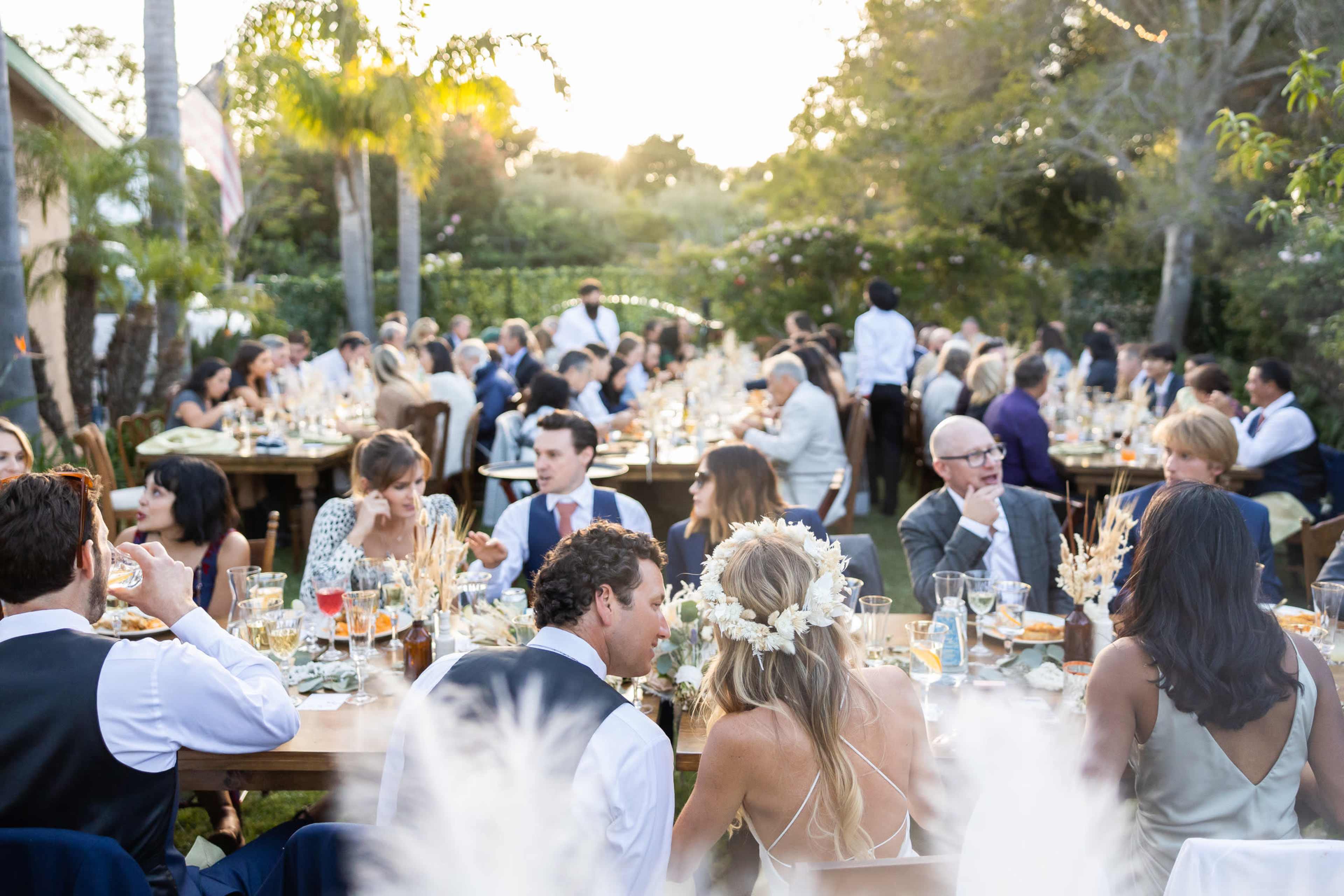 A large outdoor dinner party is set up with guests seated at long tables, enjoying their meals in a garden adorned with trees and soft lighting.