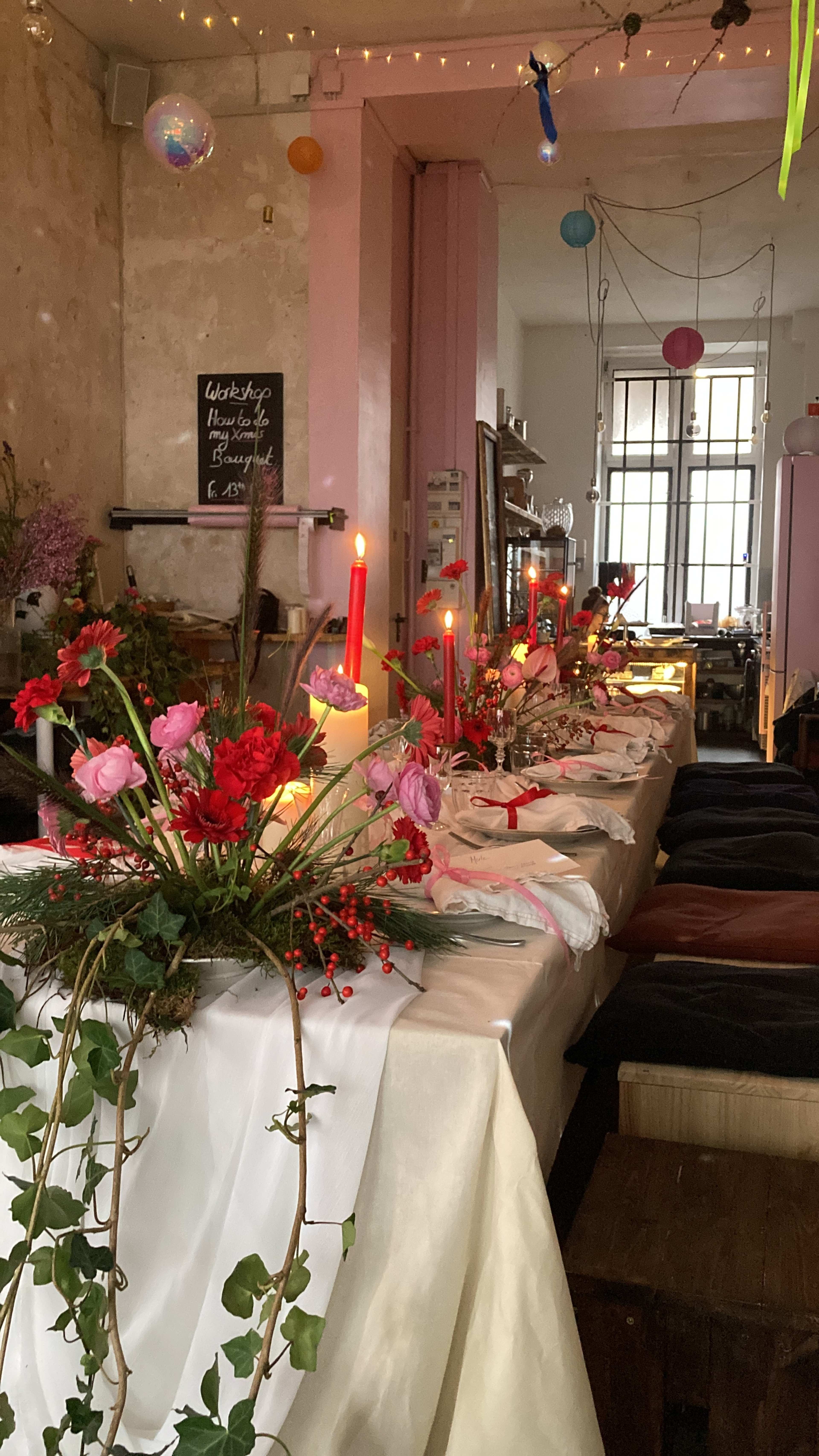 The image shows a dining table adorned with floral arrangements and candles, set in a warmly lit, rustic interior with pink walls.