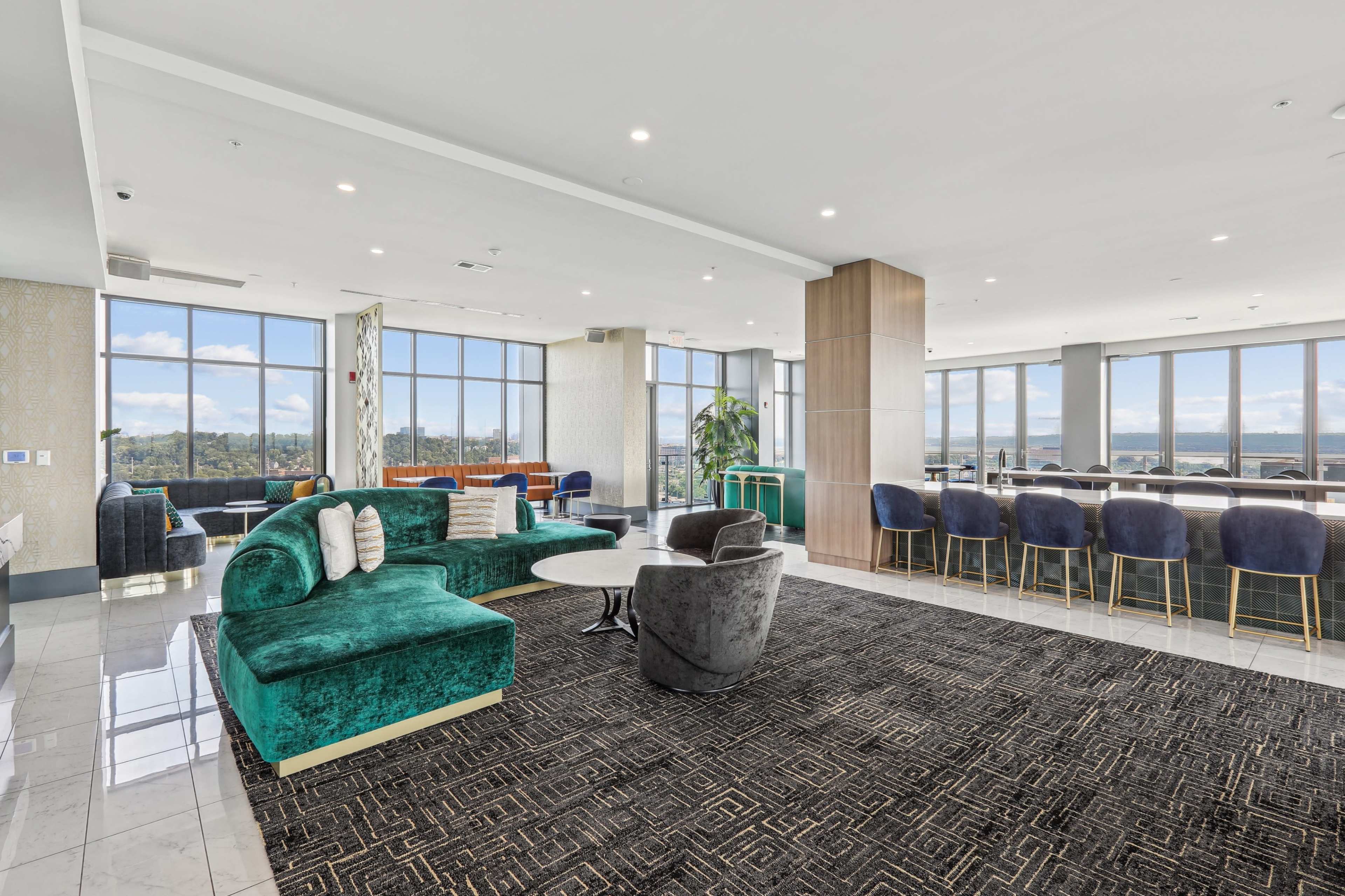 A modern lounge area with large windows, featuring a green couch, gray chairs, a coffee table, and a bar counter with high stools.