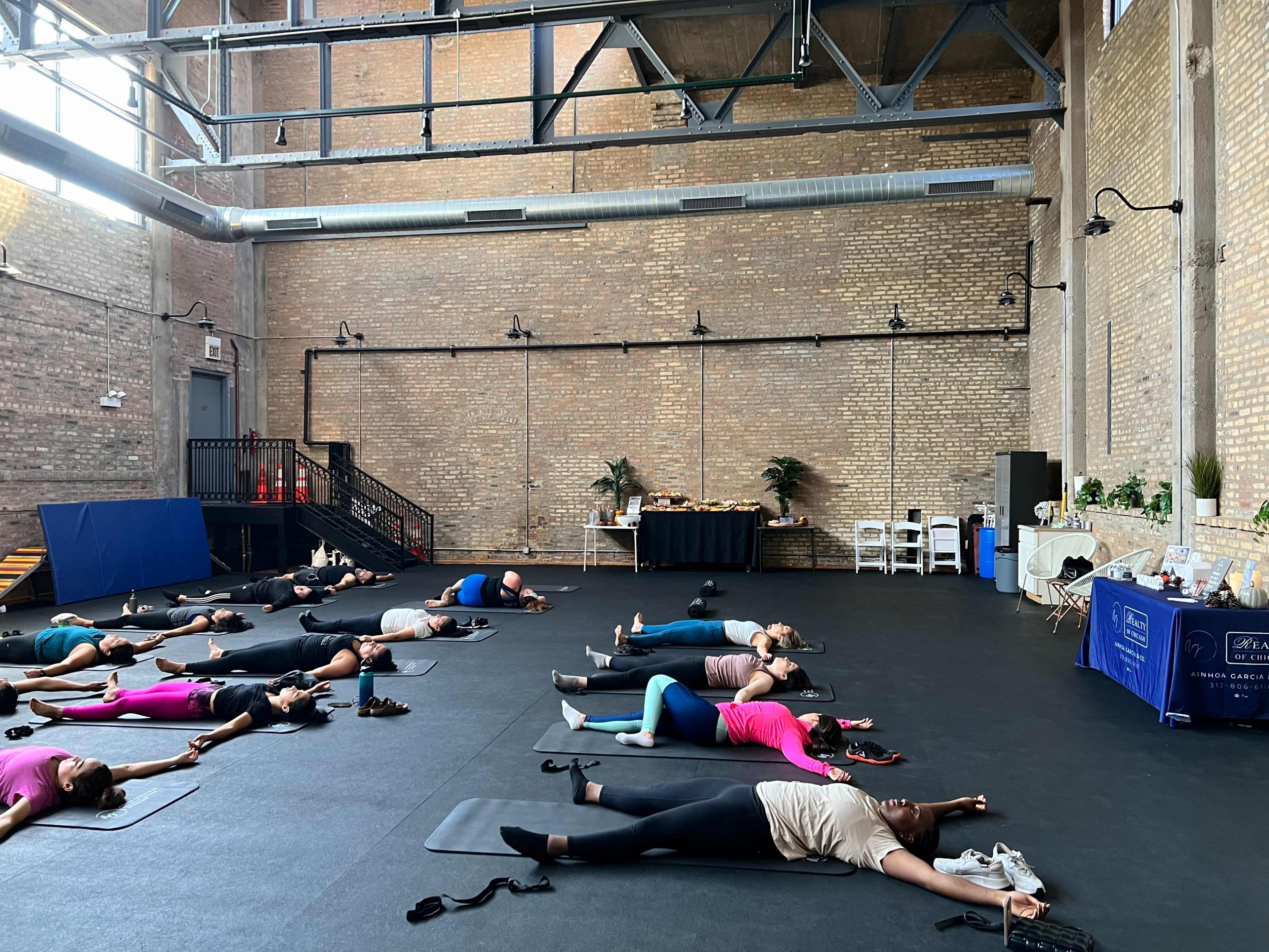 A group of people in various colored clothing practice yoga on mats in a spacious, industrial-style studio.