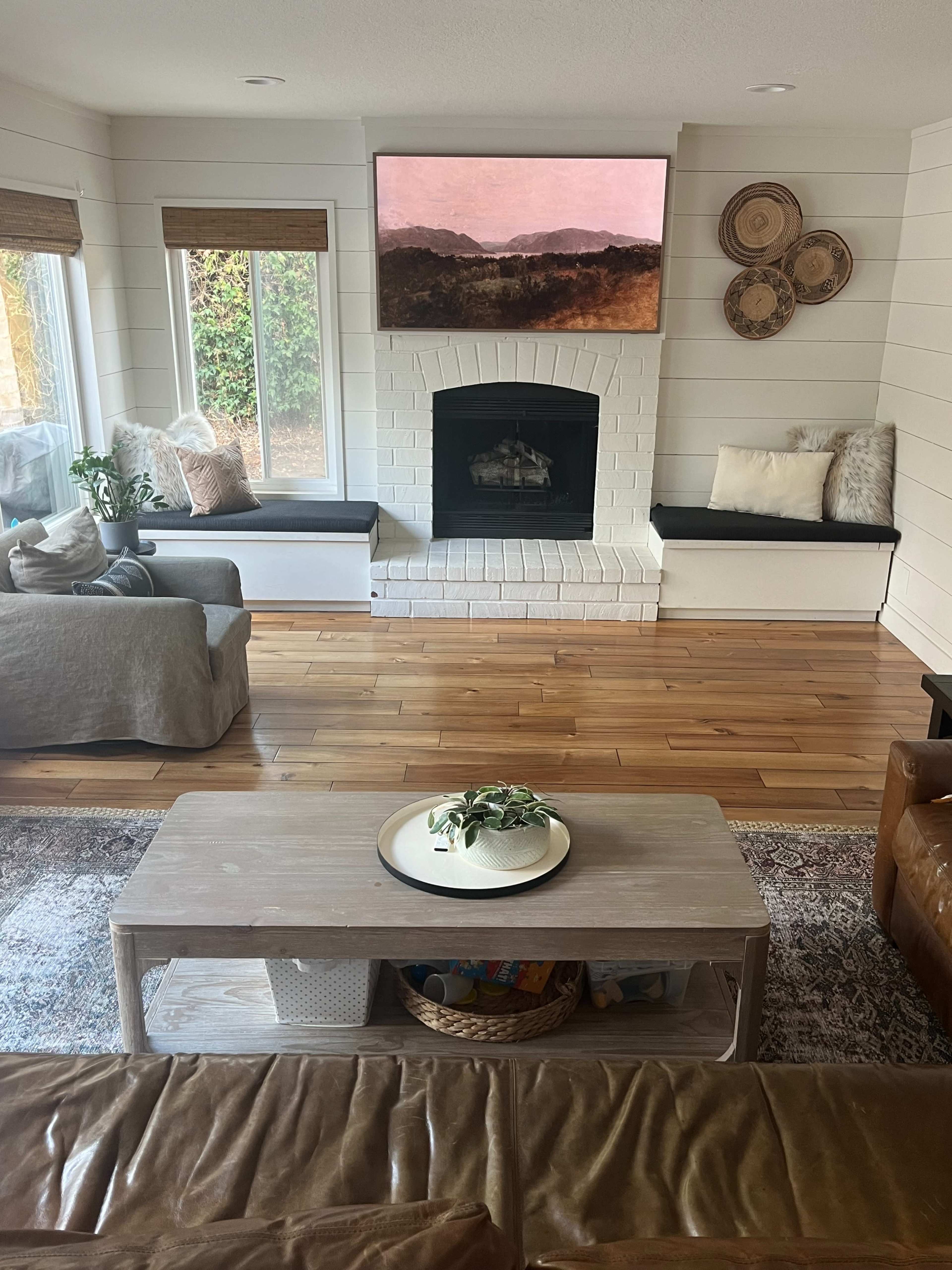 The image shows a living room featuring a white brick fireplace, large windows with natural light, and a coffee table in front of comfortable seating.