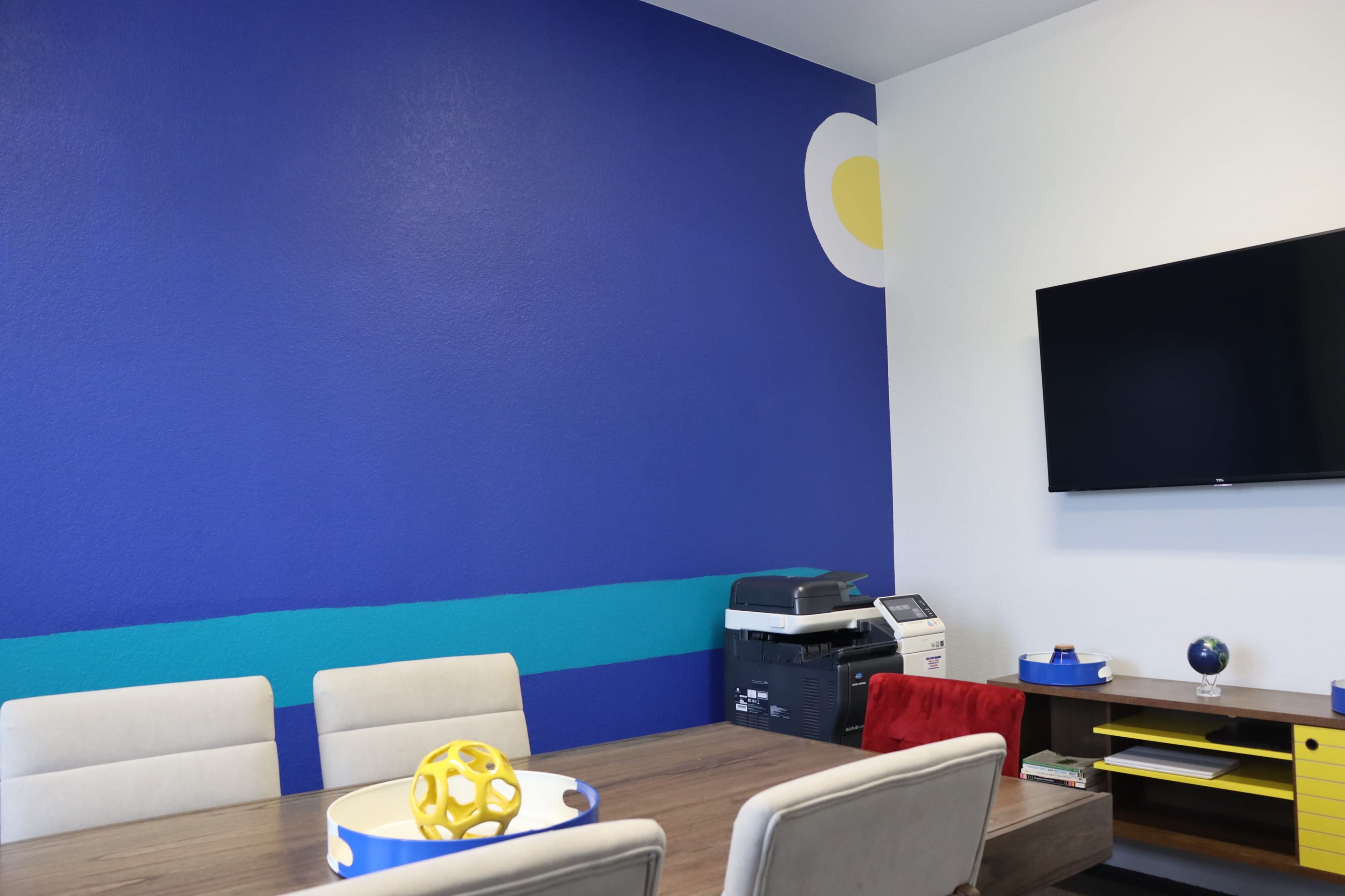 A conference room features a blue accent wall with a yellow circle, a wooden table with beige chairs, and a wall-mounted television.