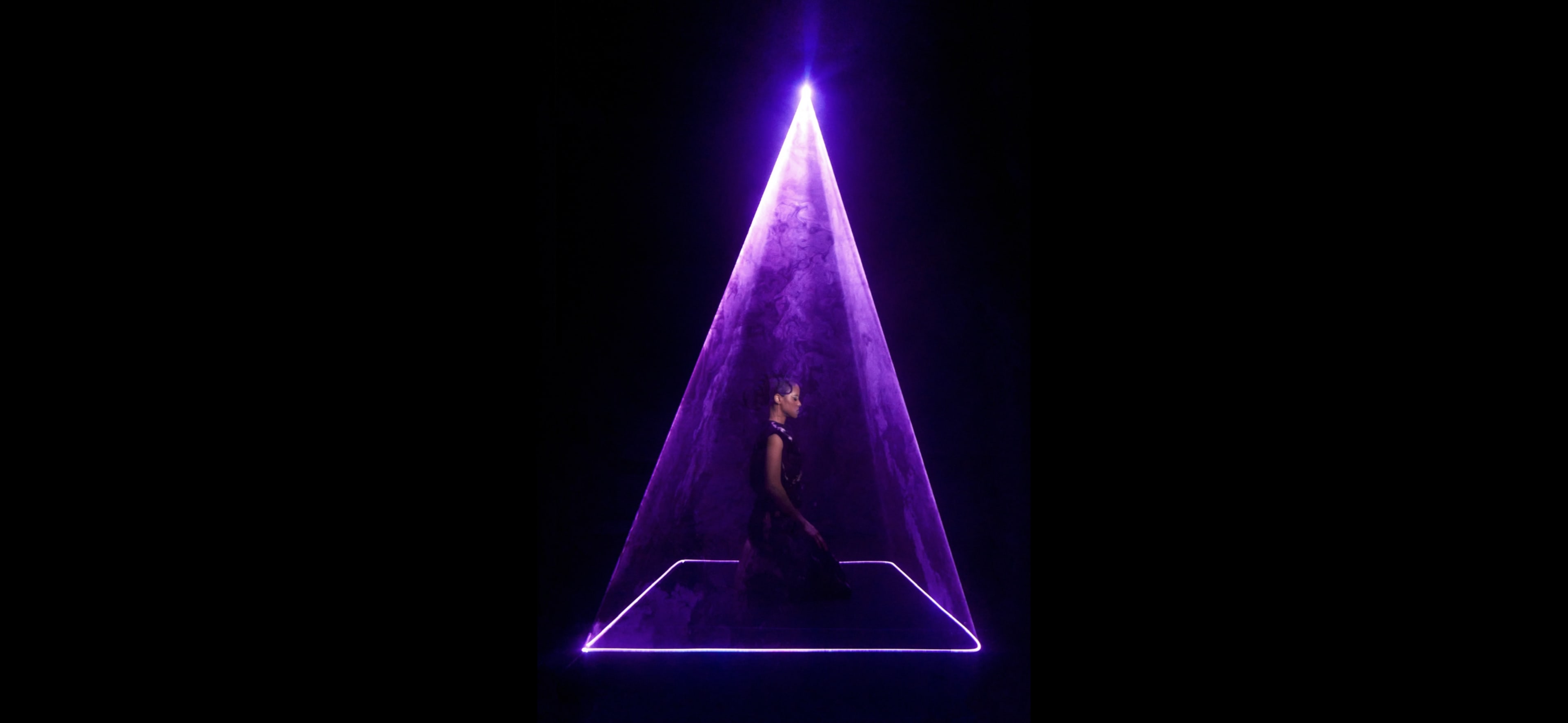A person is seated inside a pyramid-shaped structure illuminated by purple light and surrounded by smoke.