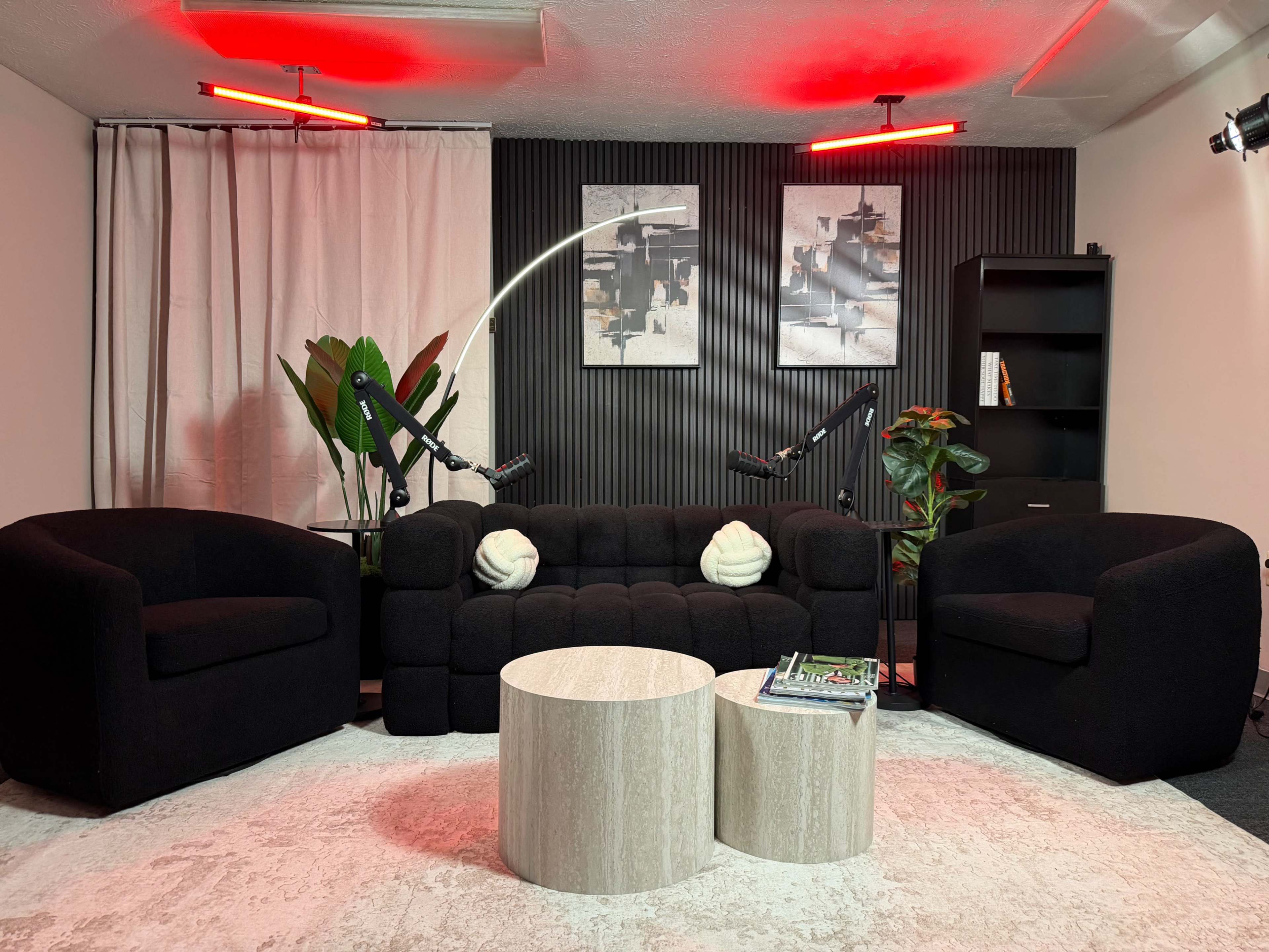 The image shows a modern, black lounge area with two curved sofas, a pair of round tables, potted plants, and artwork on the walls, illuminated by red overhead lights.