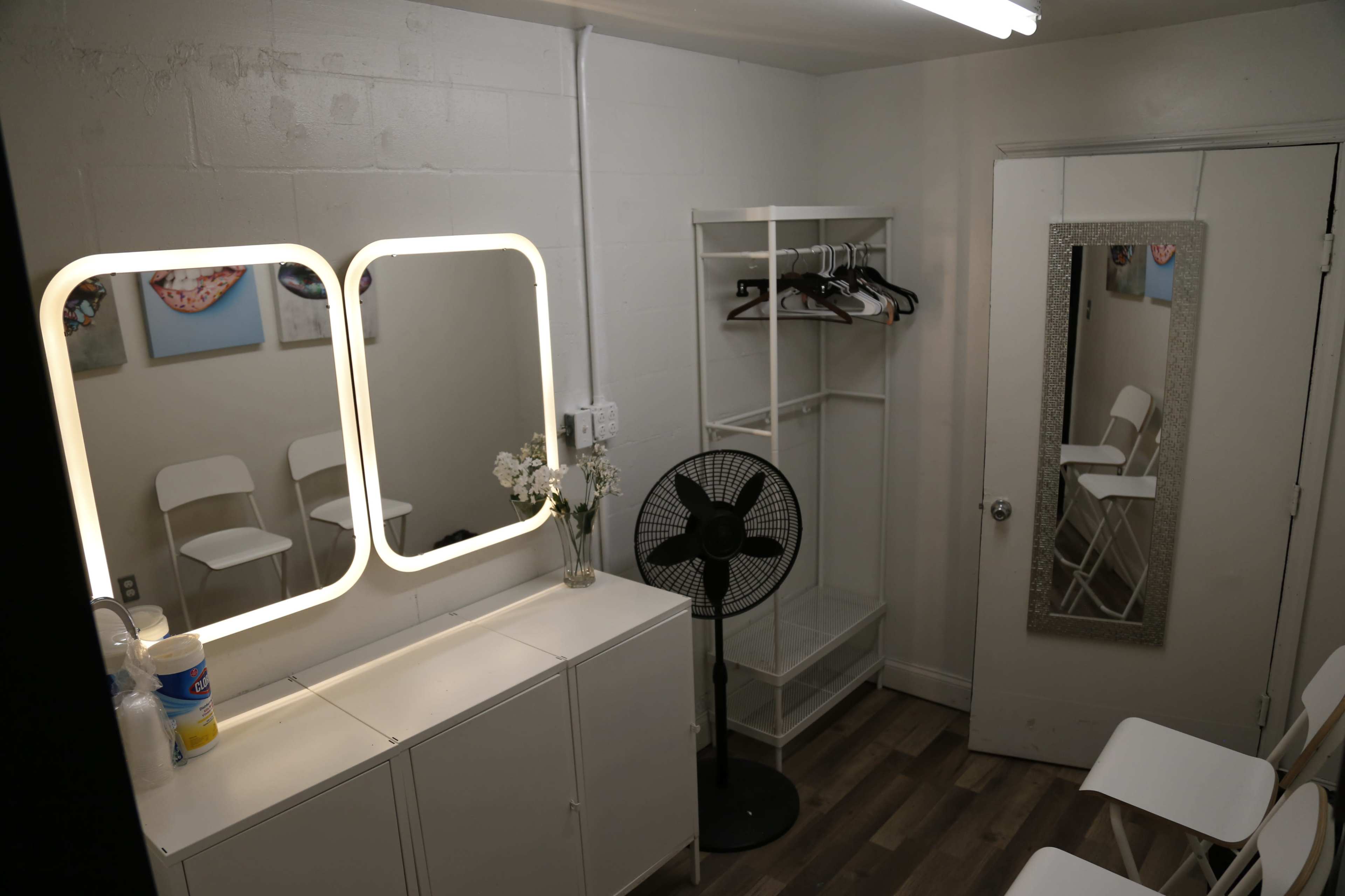A simple room with two illuminated mirrors, a fan, a clothing rack, and white folding chairs arranged against the walls.