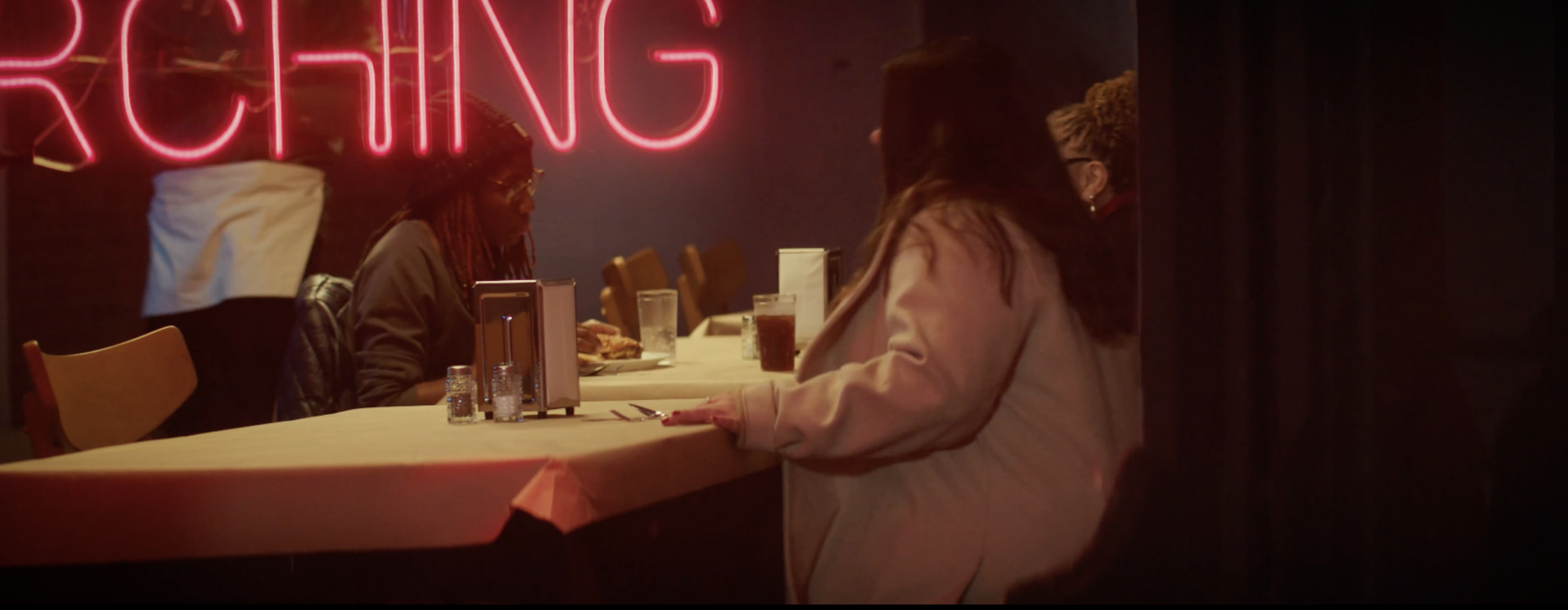 A group of people sits at a table in a dimly lit restaurant, with a neon sign reading "SEARCHING" glowing in the background.