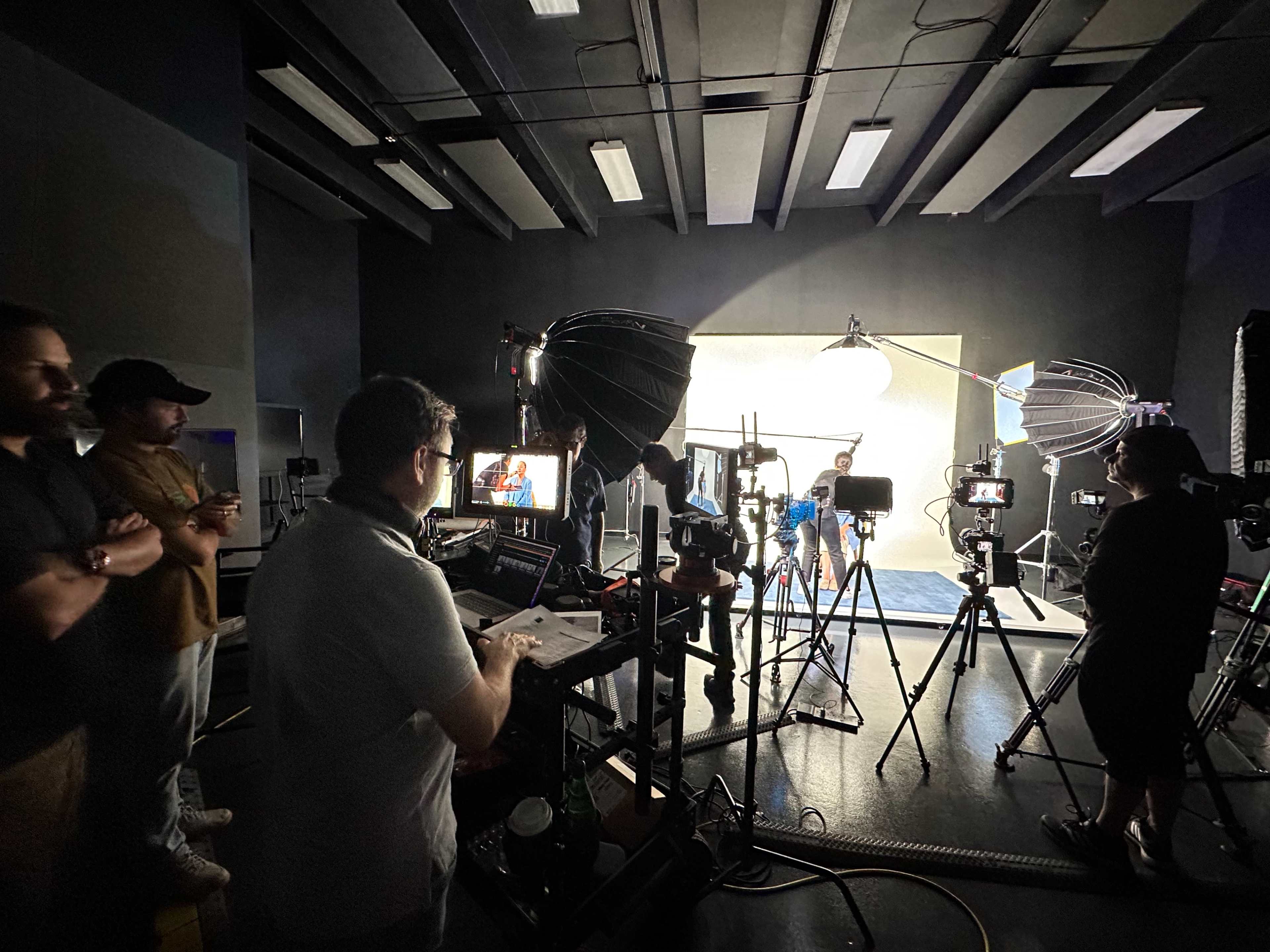 A film crew sets up lighting and equipment in a studio while a model poses against a backdrop.