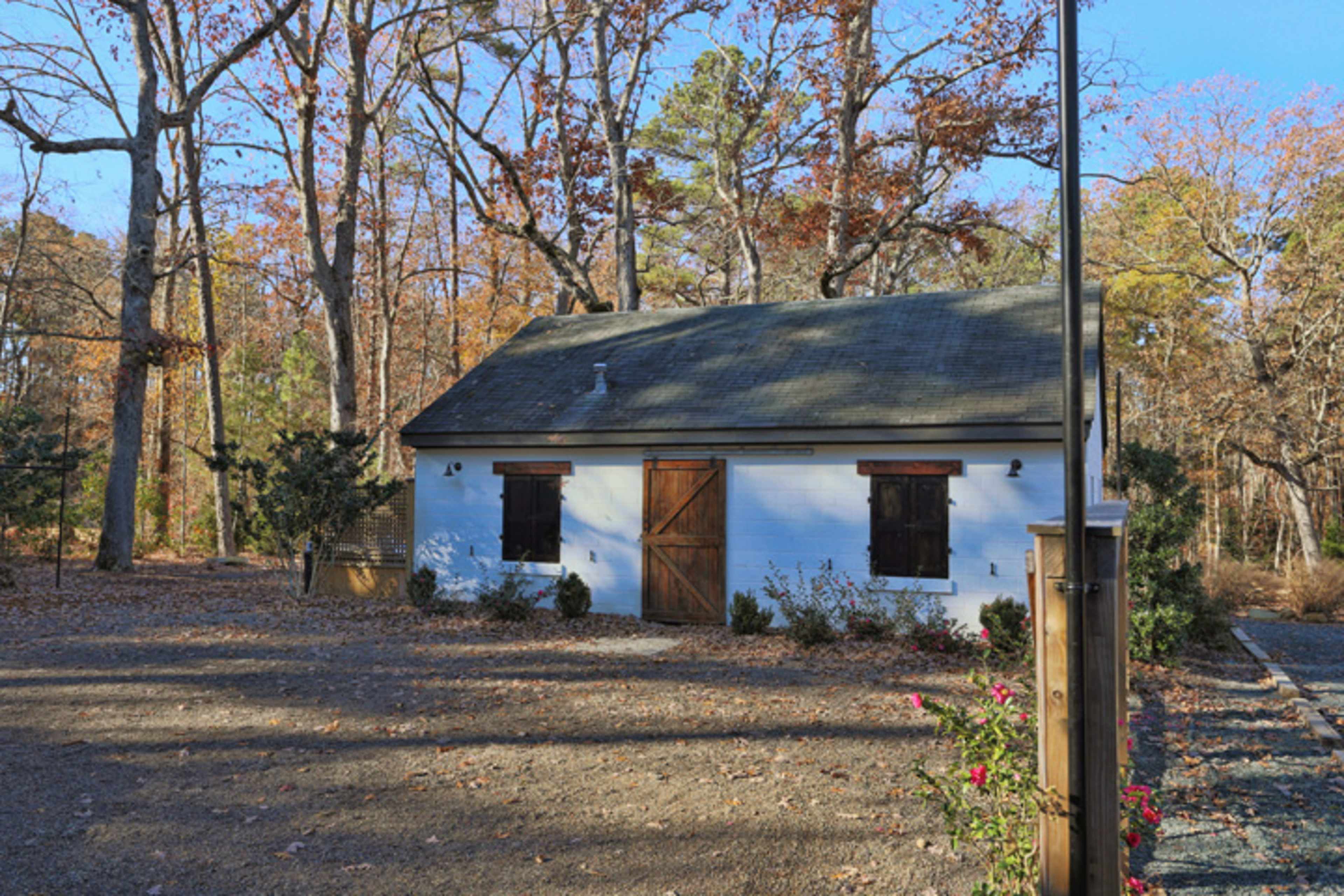 A small white cabin with brown shutters and a wooden door, surrounded by trees shedding autumn leaves.