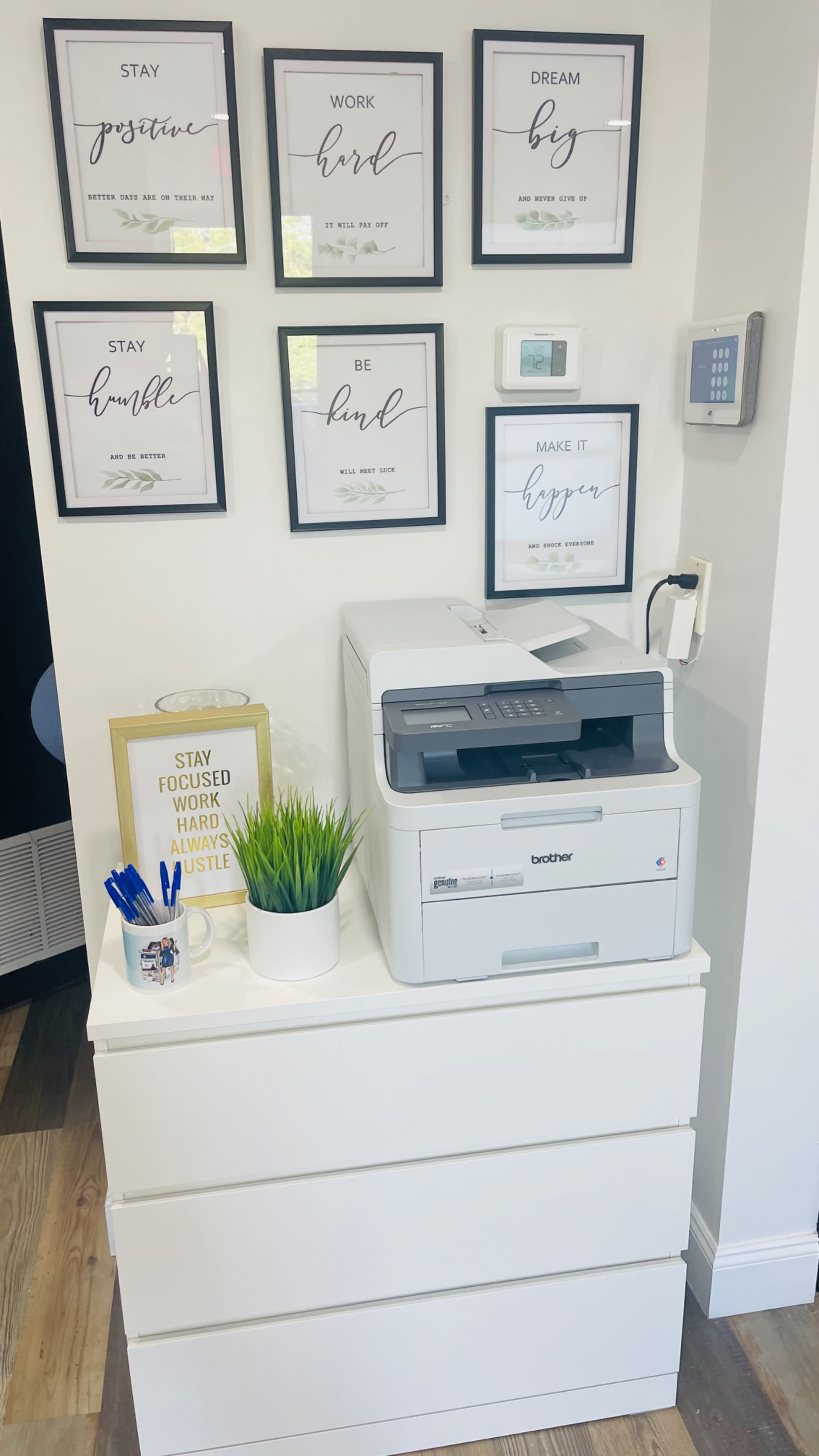 A modern office corner features a printer on a white drawer unit, surrounded by framed motivational quotes and a small decorative plant.
