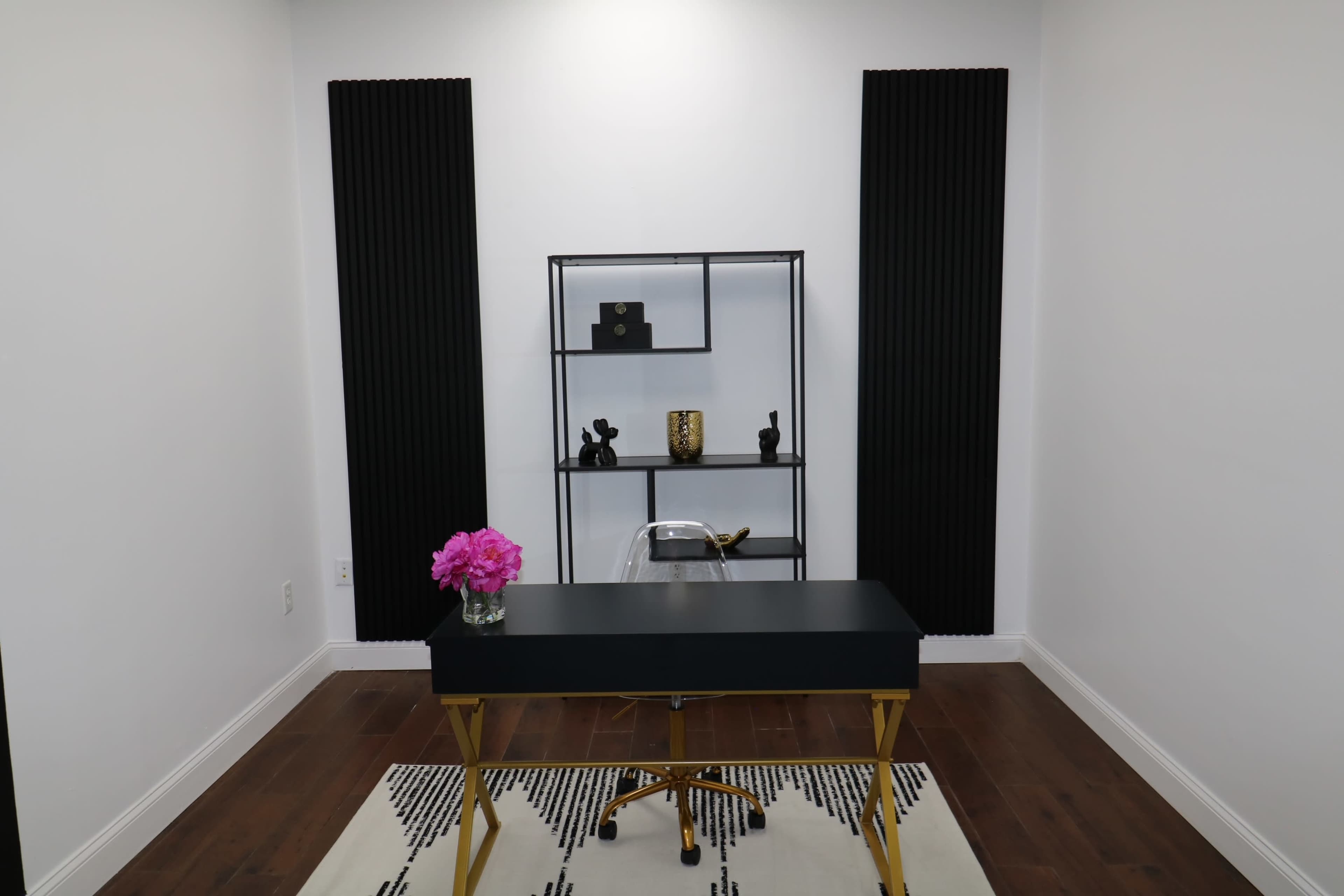 A modern office space featuring a black desk with gold legs, a decorative shelving unit against the white walls, and a small floral arrangement in front.