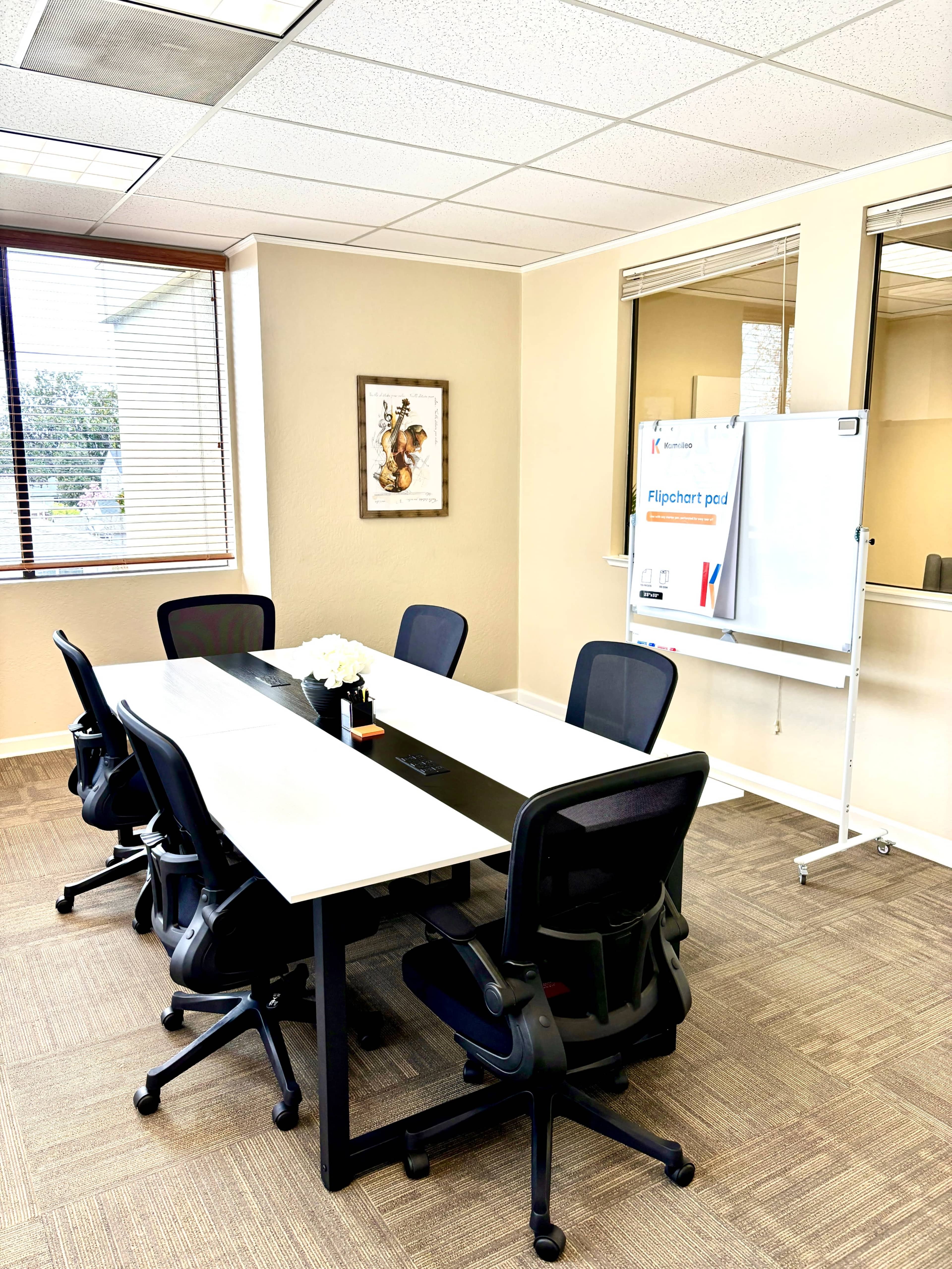 A conference room with a long white table surrounded by six black chairs, a flower arrangement in the center, and a flipchart stand nearby.