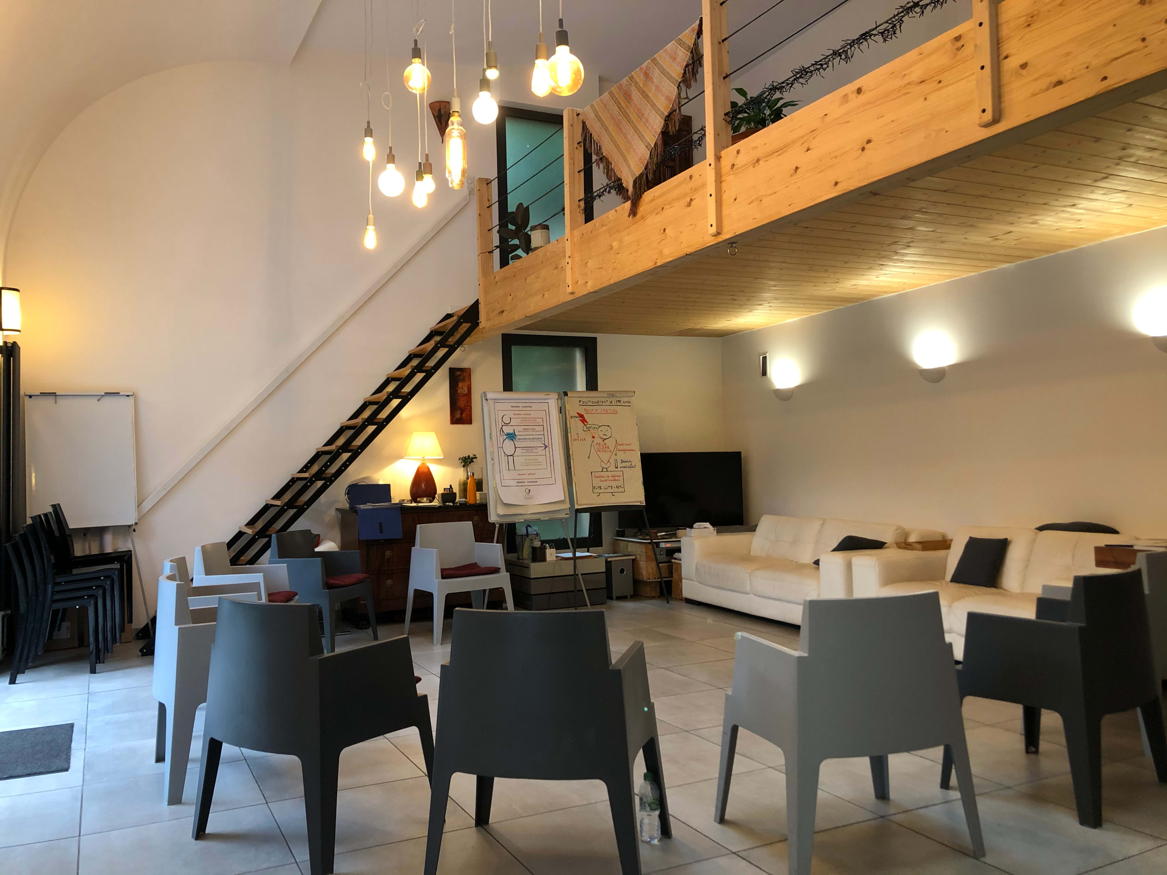 An interior room with a seating area arranged in a circle, a staircase leading to a wooden loft, and a presentation board beside a television.