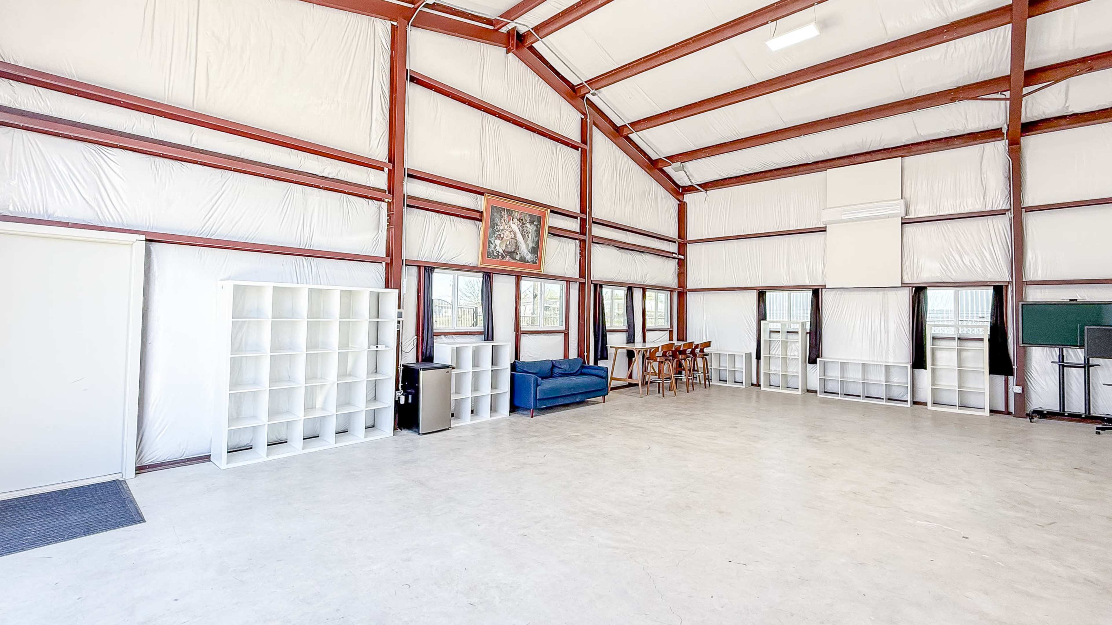 The image shows a spacious, indoor area with high ceilings, featuring metal beams, a blue couch, a table with chairs, and white shelves lining the walls.