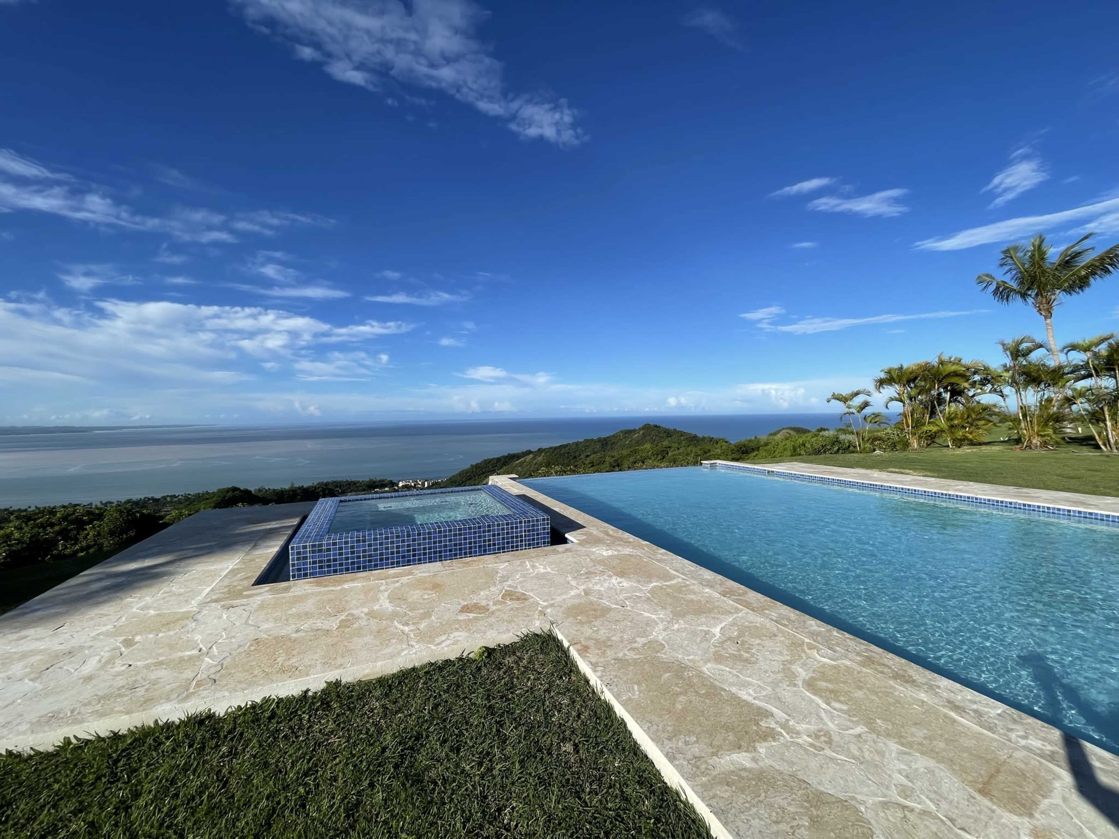 A modern infinity pool with a view overlooking the ocean and surrounding hills is situated against a clear blue sky.
