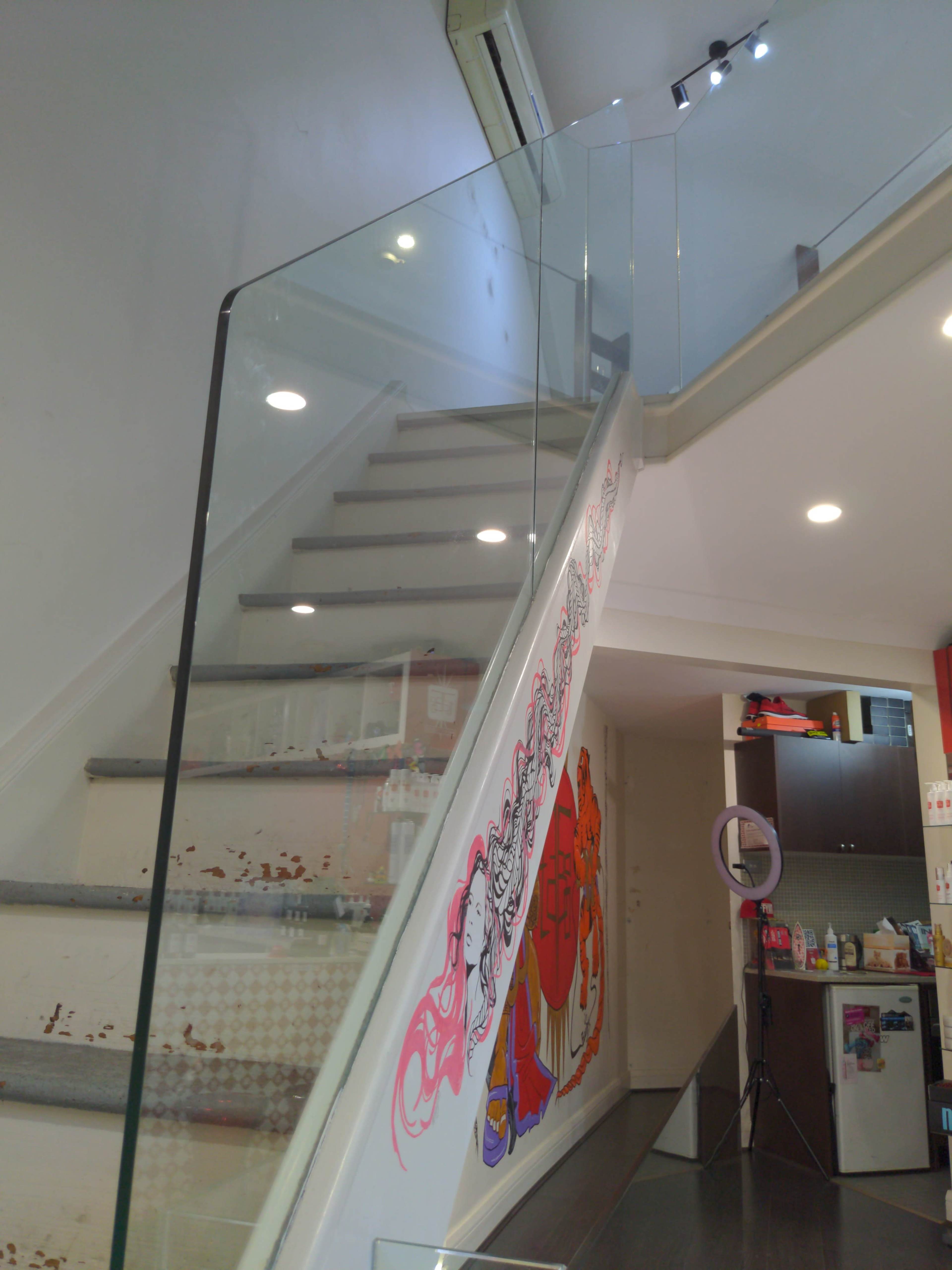 The image shows a glass-railed staircase leading to an upper level, with colorful artwork on the wall and a well-lit space at the top.