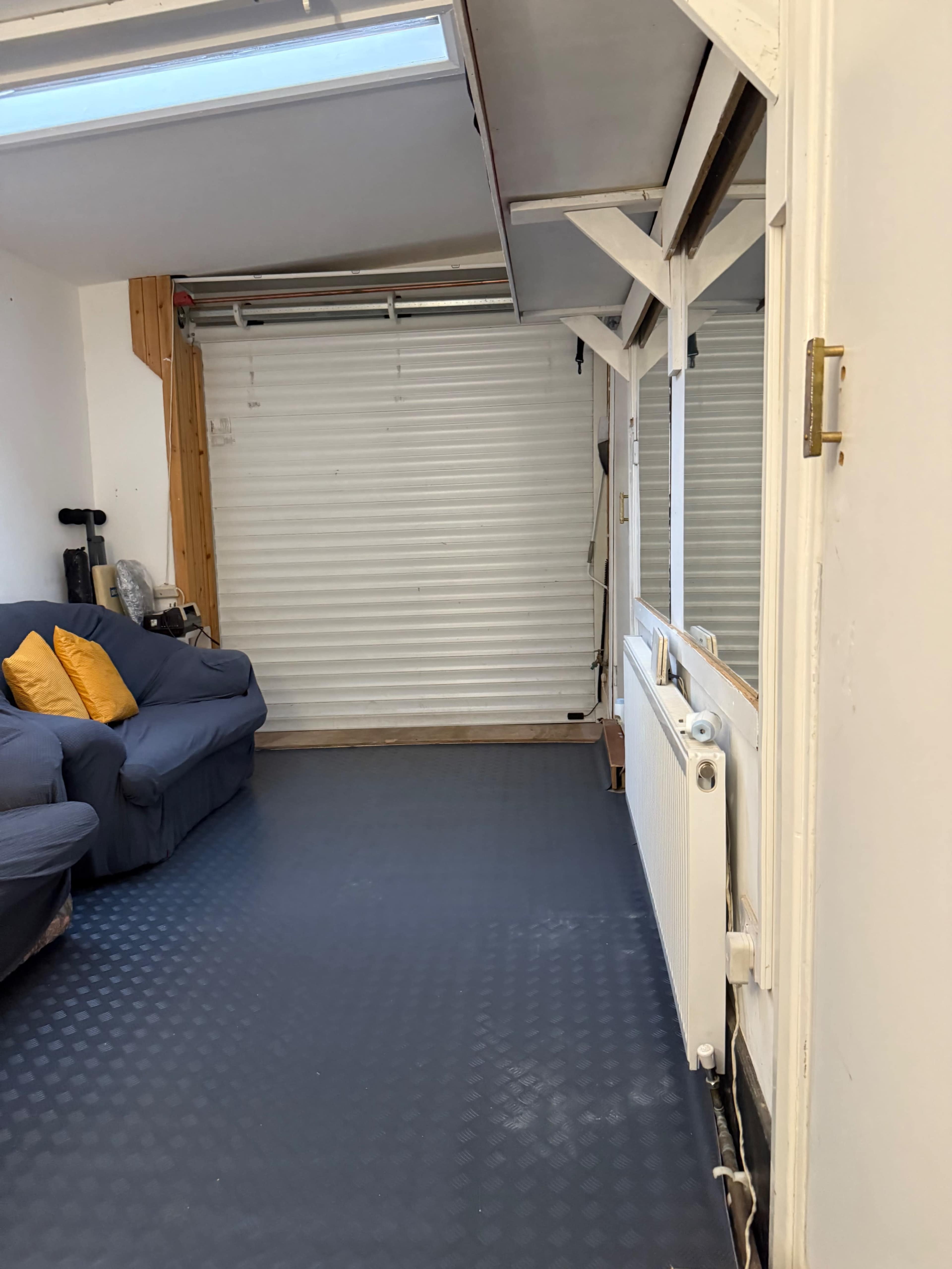 The image shows a room with a blue sofa facing a wall, a radiator on the side, and a roll-up door at the end of the space.