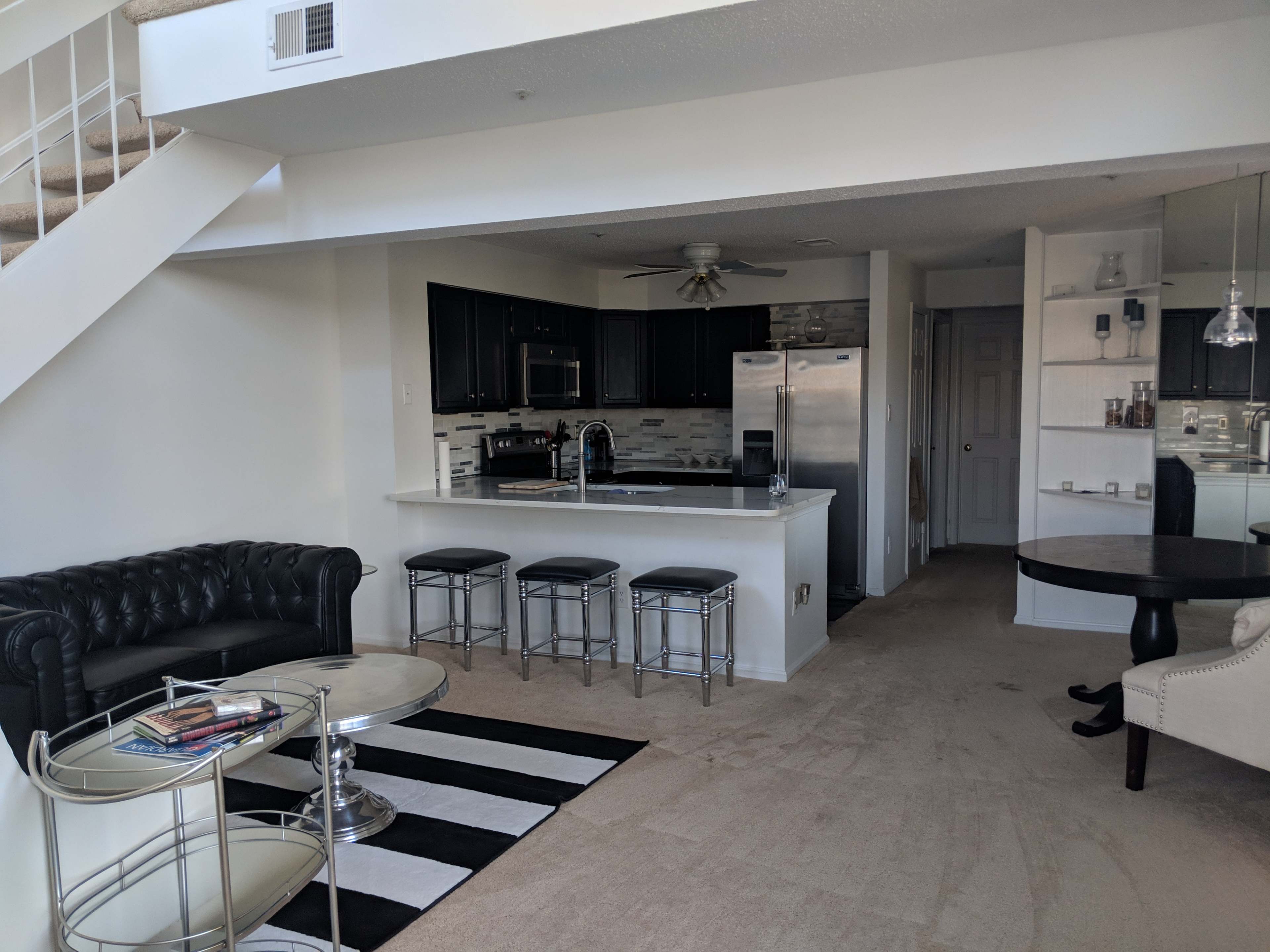 A modern, open-layout living space featuring a kitchen with dark cabinetry, stainless steel appliances, and a living area with a black sectional couch and glass coffee tables on a striped rug.